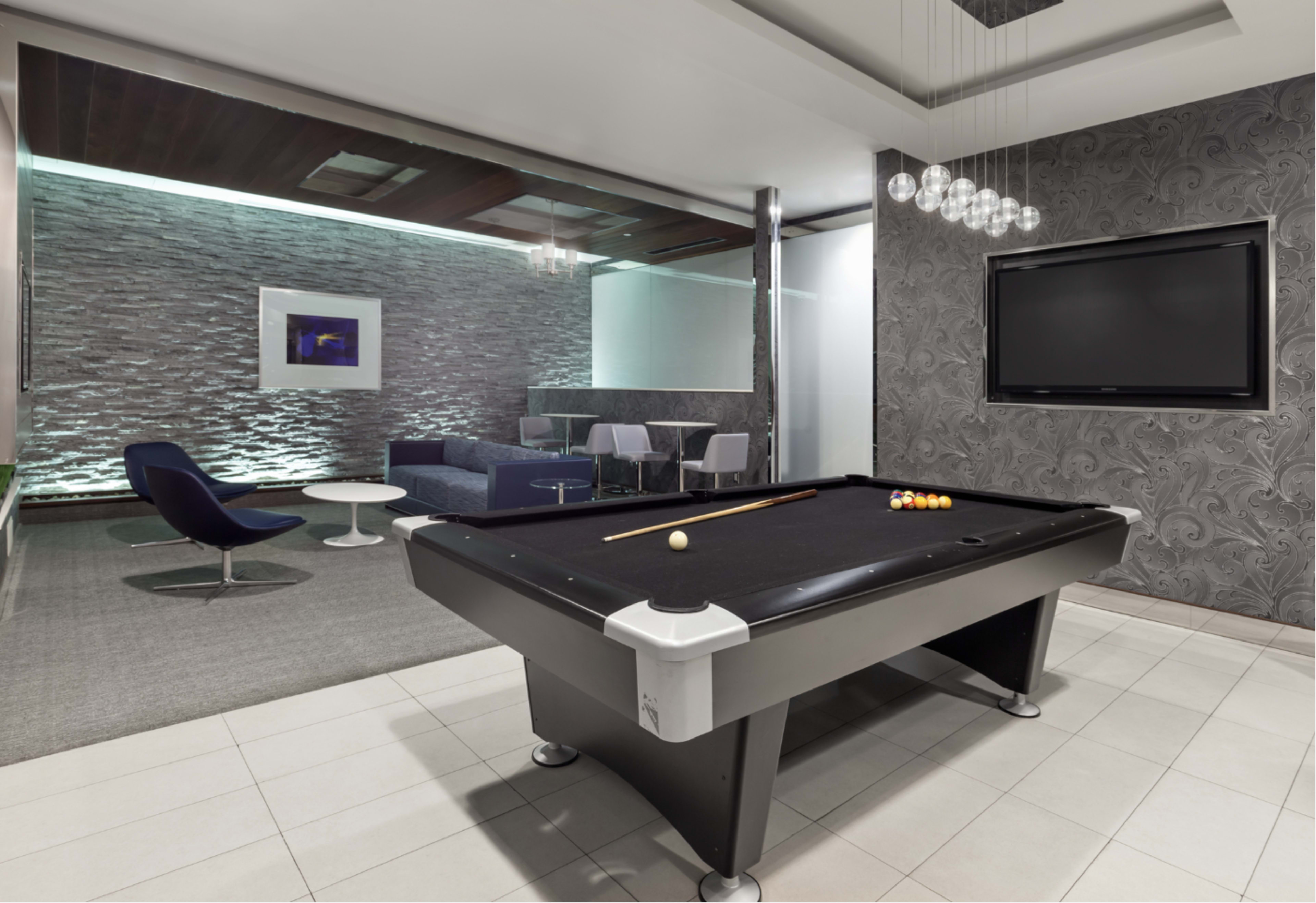 The image shows a modern game room featuring a black pool table, decorative wall panels, and a seating area with contemporary furniture.
