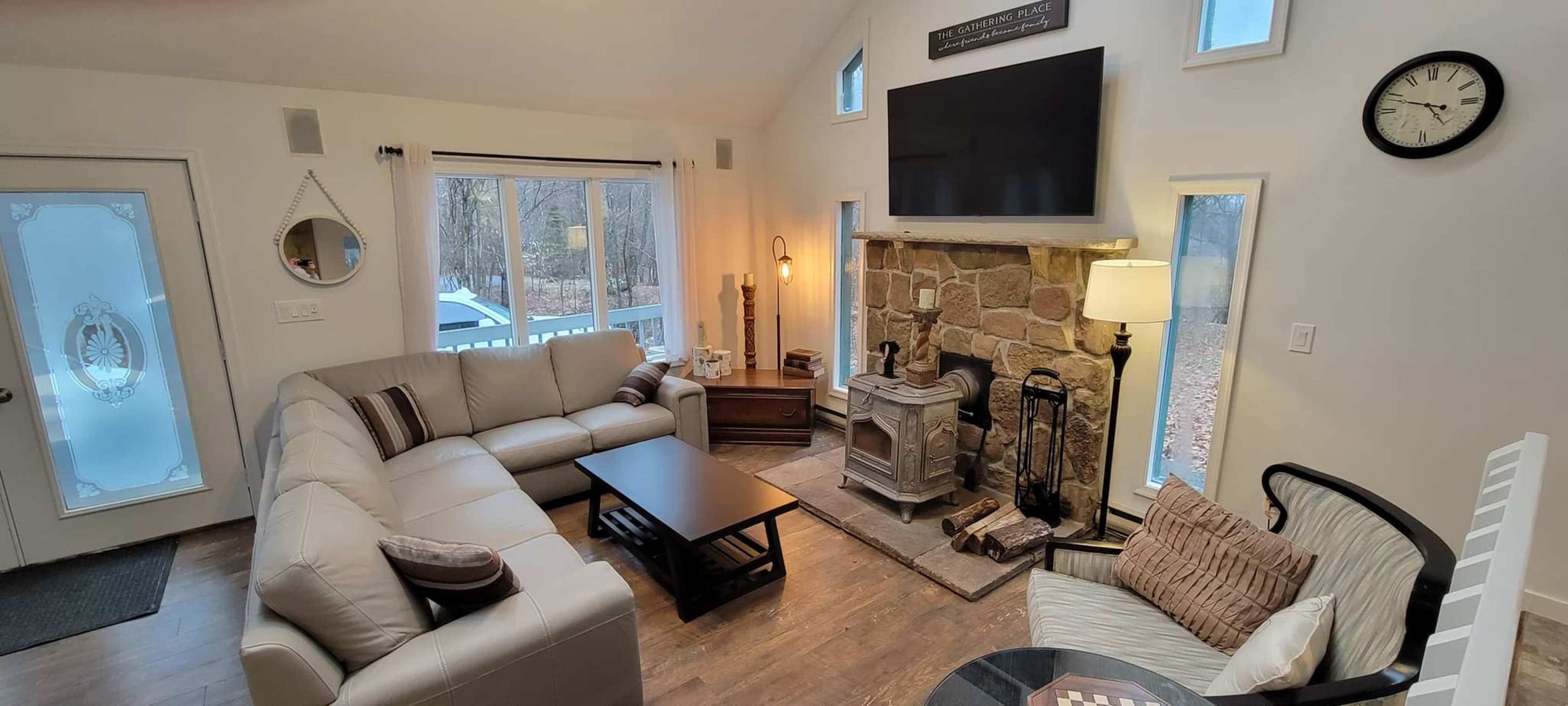 The image shows a living room with a light-colored sectional sofa, a coffee table, a stone fireplace with a wood stove, and large windows providing natural light.