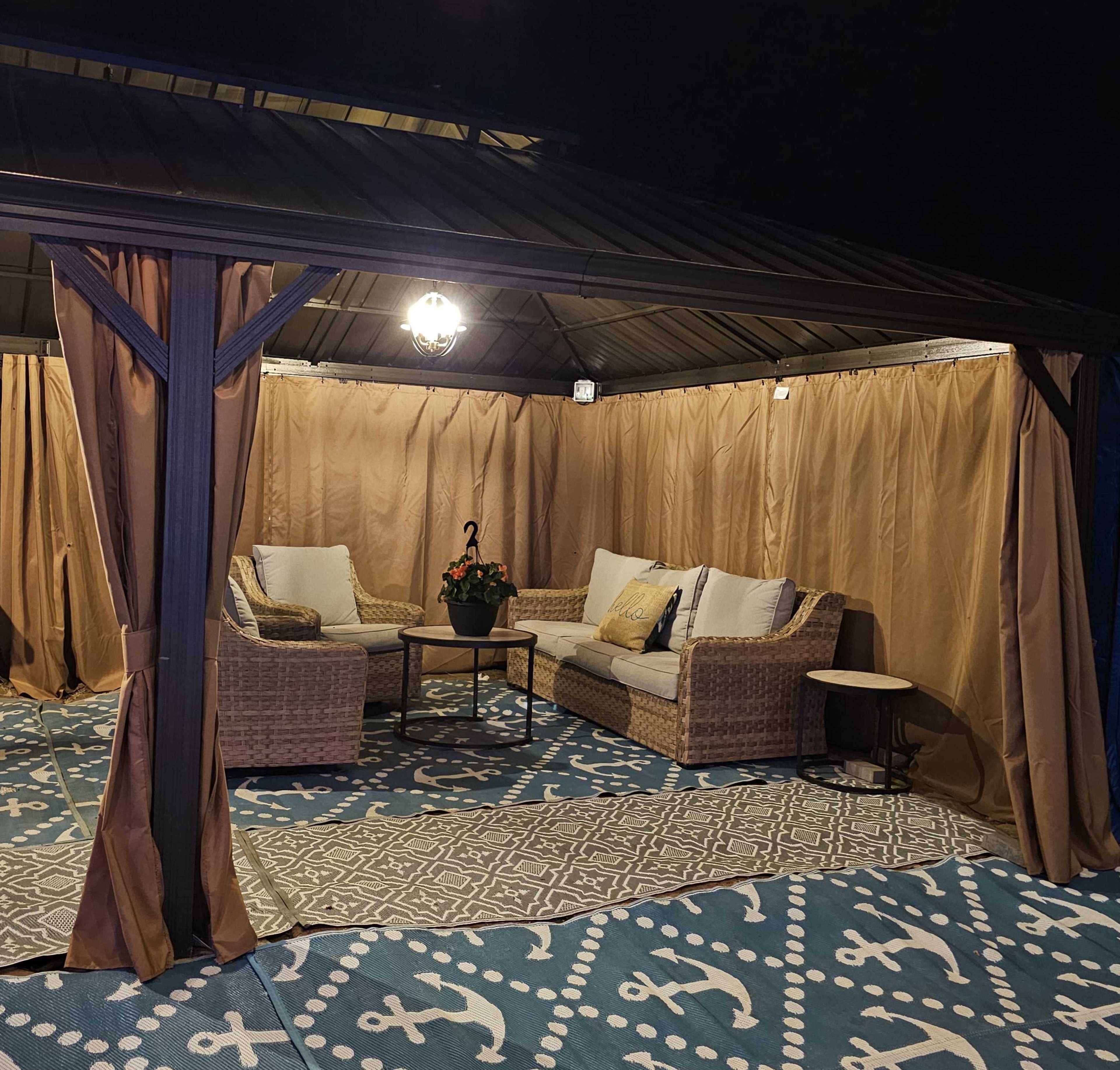 A gazebo with draped fabric contains a seating area with two chairs, a small table, and a potted plant on a patterned rug.