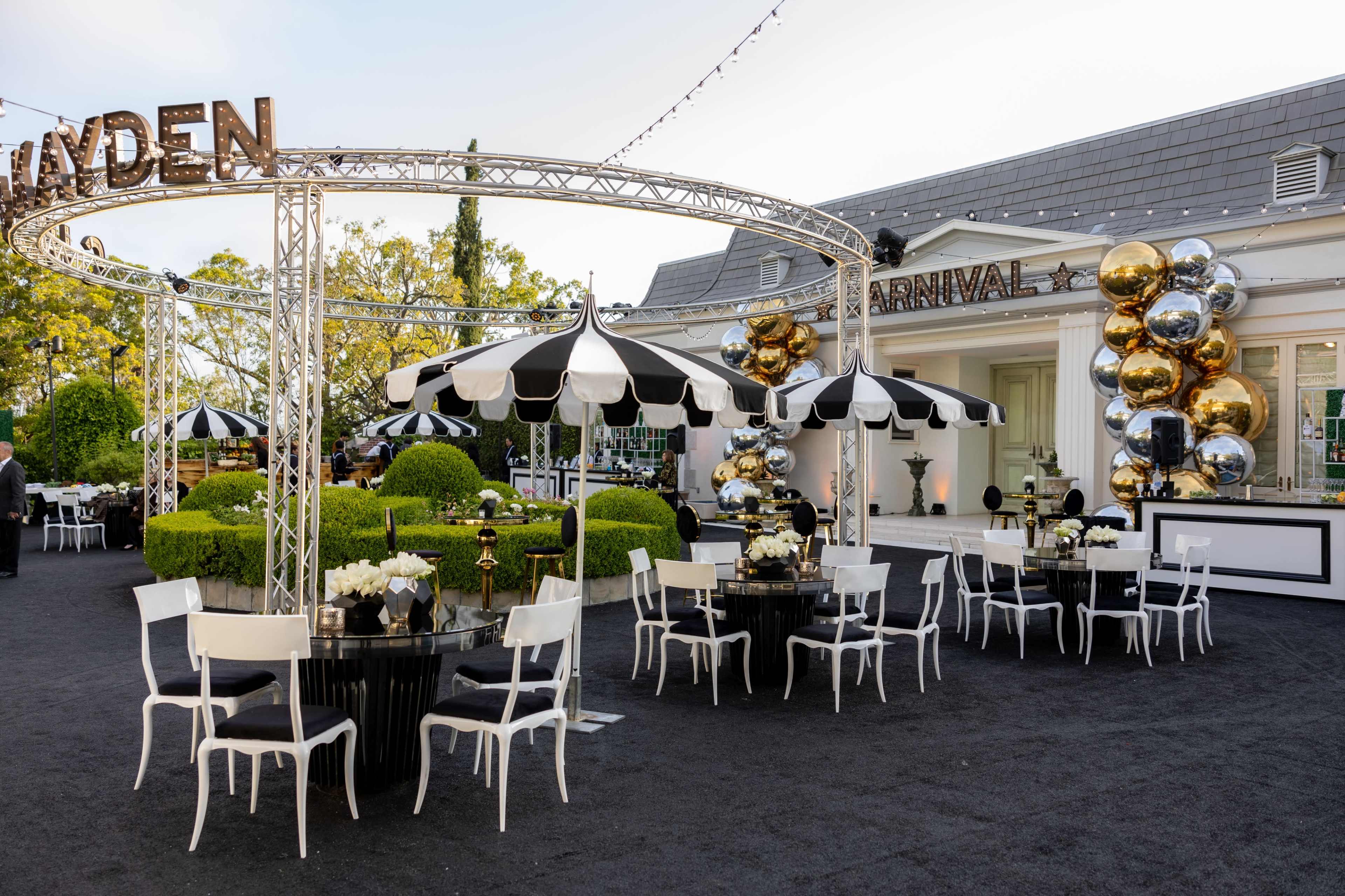 An outdoor event space featuring black and white striped umbrellas, circular tables adorned with white floral arrangements, and a backdrop of a building decorated with balloons.