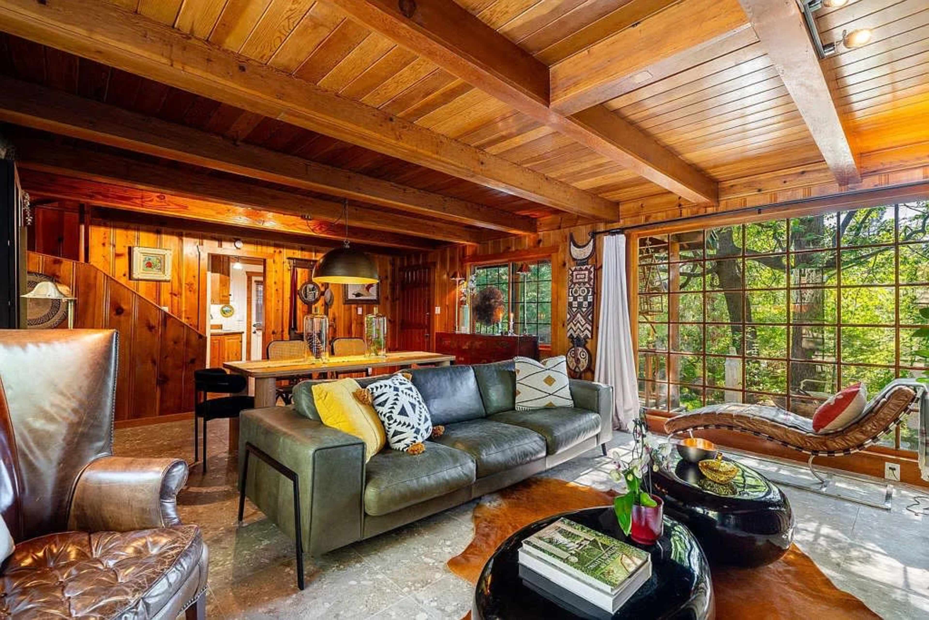 The image shows a cozy living room featuring wooden beams, a leather couch, a dining area, and large windows overlooking greenery.