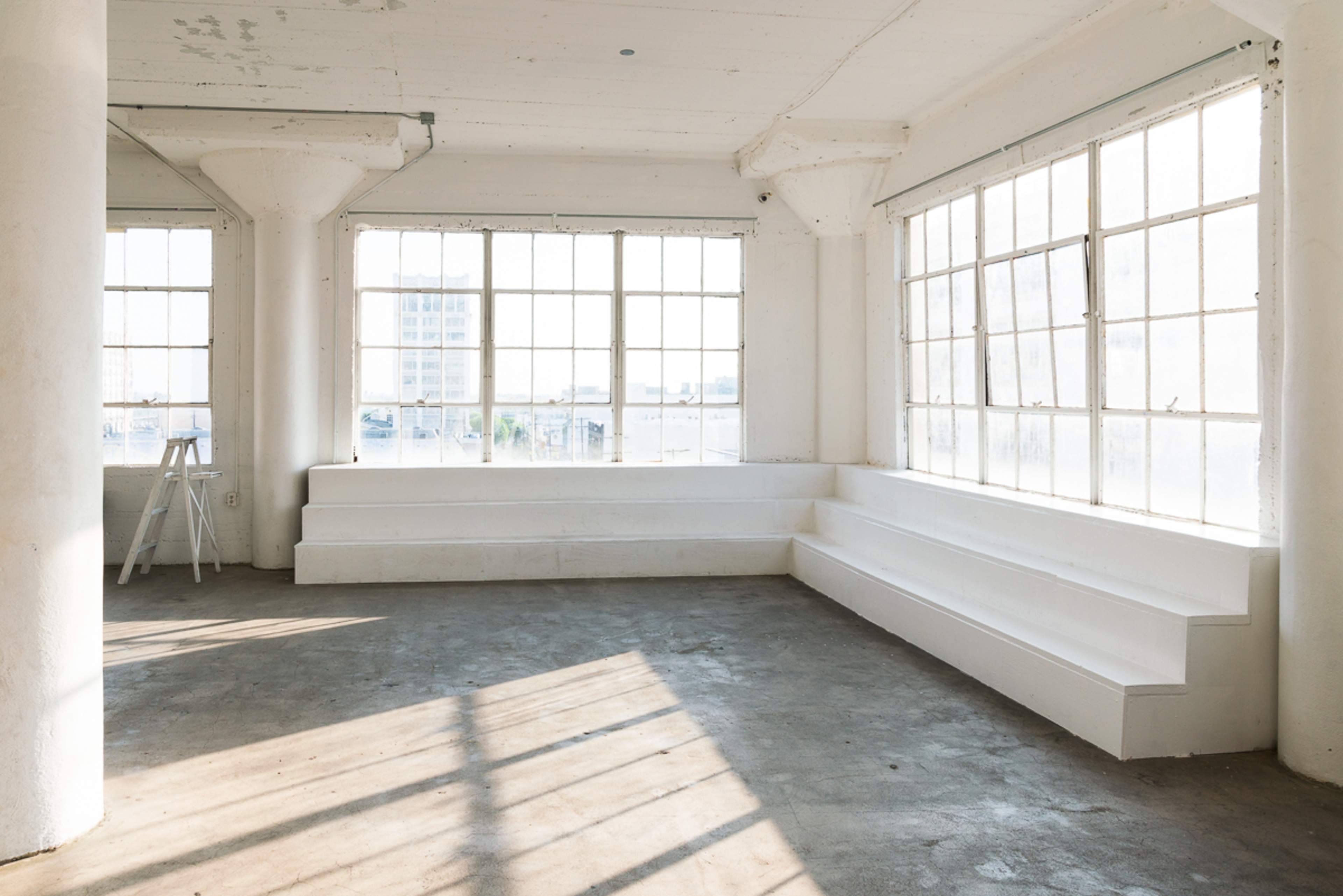 A bright, empty room with white walls and large windows, featuring built-in white seating along two sides and a concrete floor.