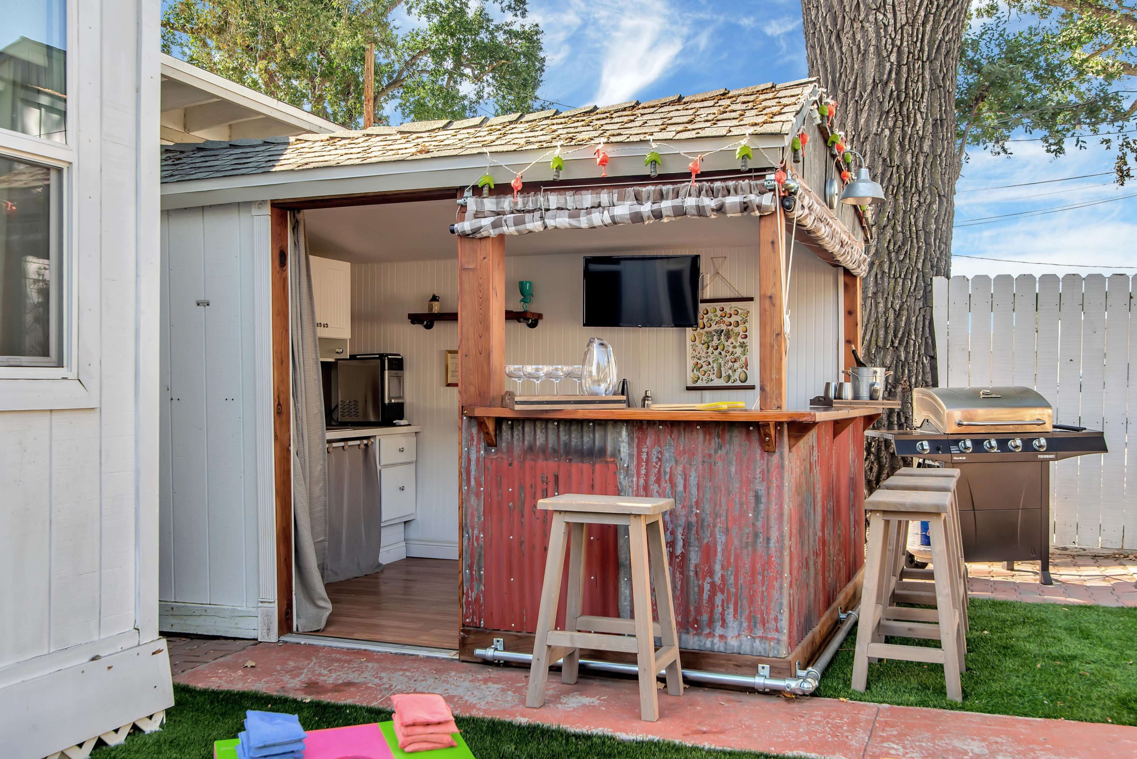A rustic outdoor bar with a red corrugated metal front, wooden stools, and a television inside, situated next to a house and a grill in a backyard setting.