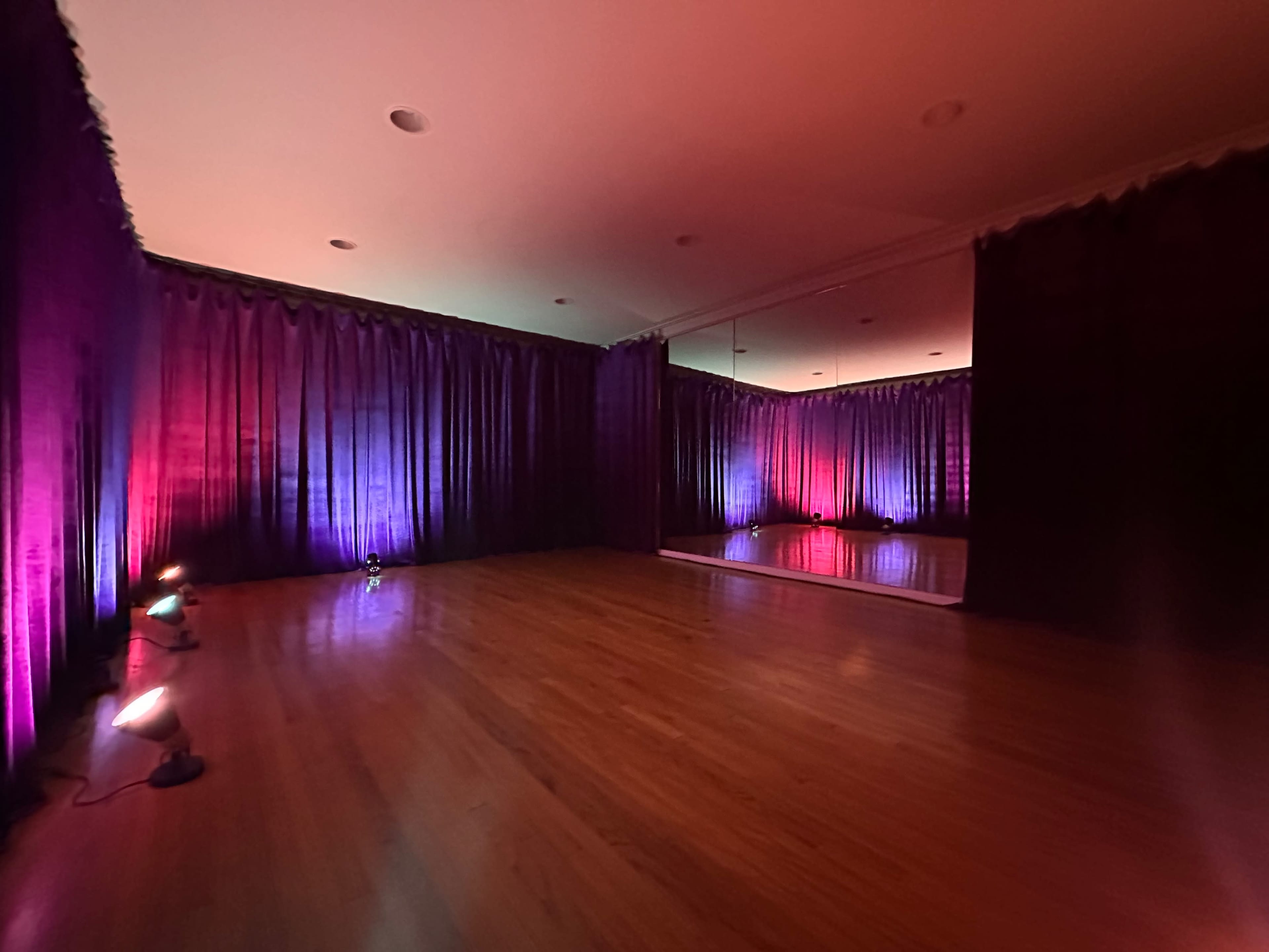 The image shows a spacious dance studio with wooden floors, mirrored walls, and colored lights creating a vibrant atmosphere.
