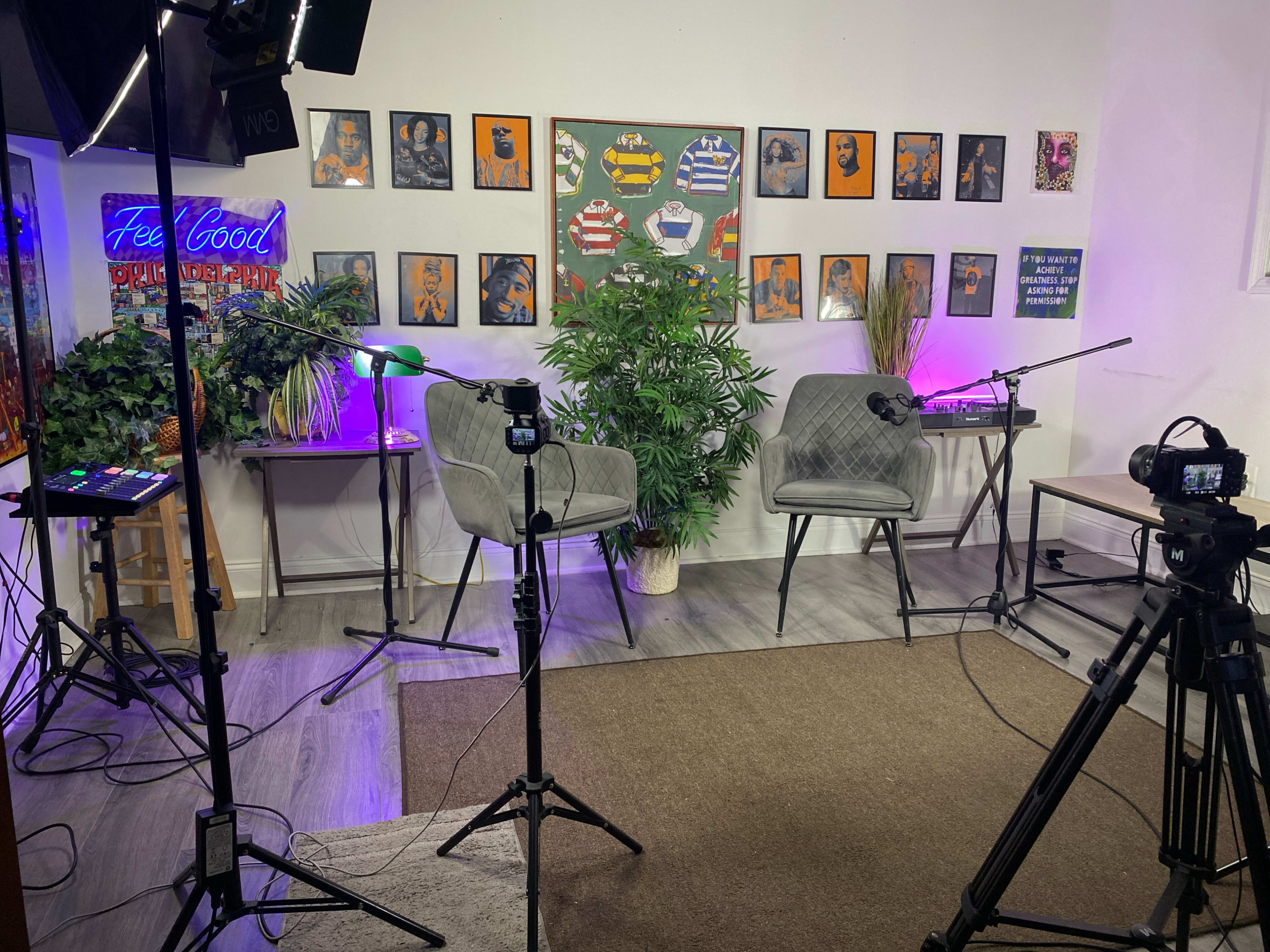 The image shows a modern talk show set with two grey chairs, a potted plant, and multiple cameras positioned around a colorful wall decorated with framed artworks.