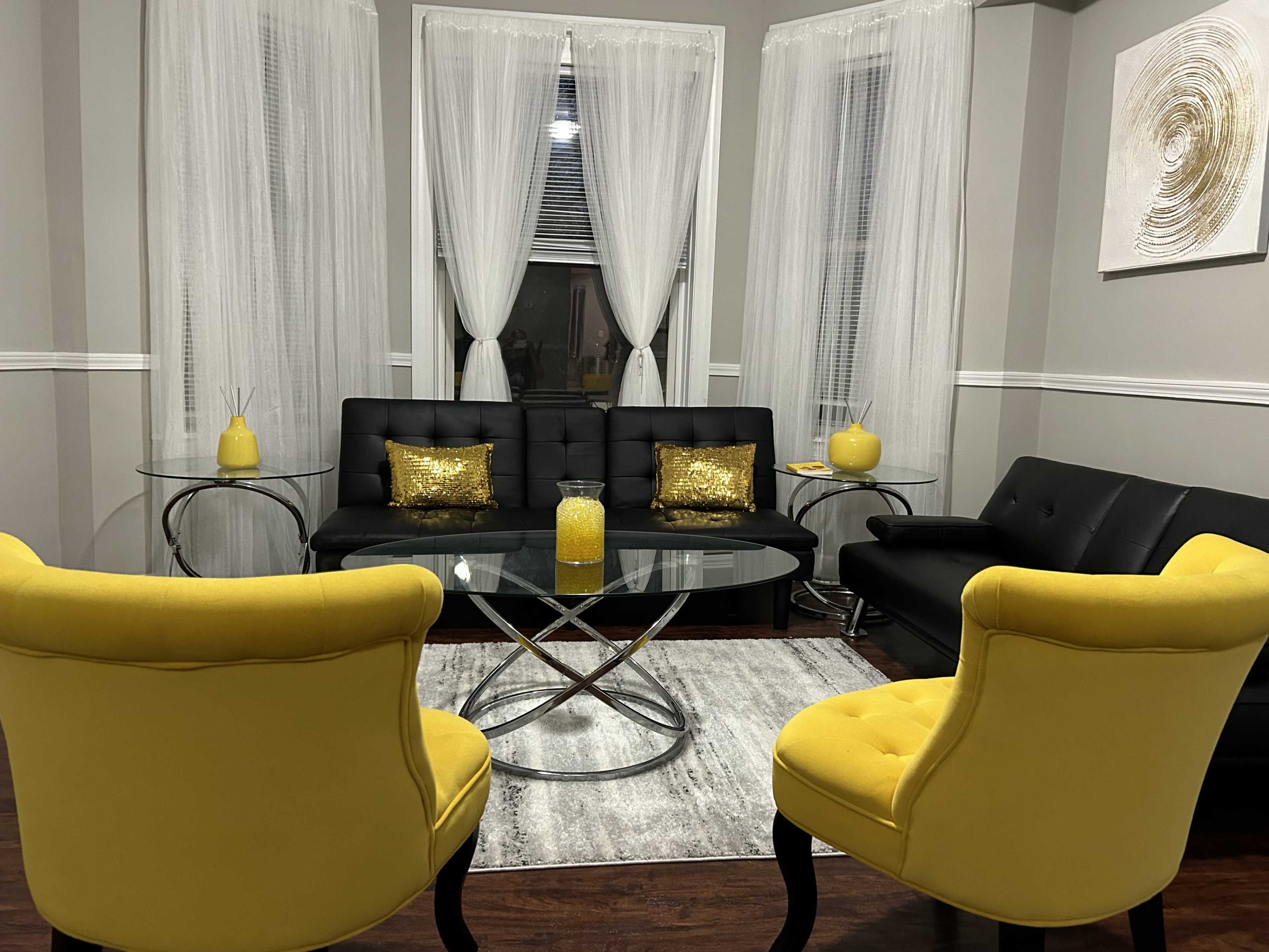 A cozy living room features a black leather couch, a glass coffee table, two yellow chairs, and sheer white curtains.