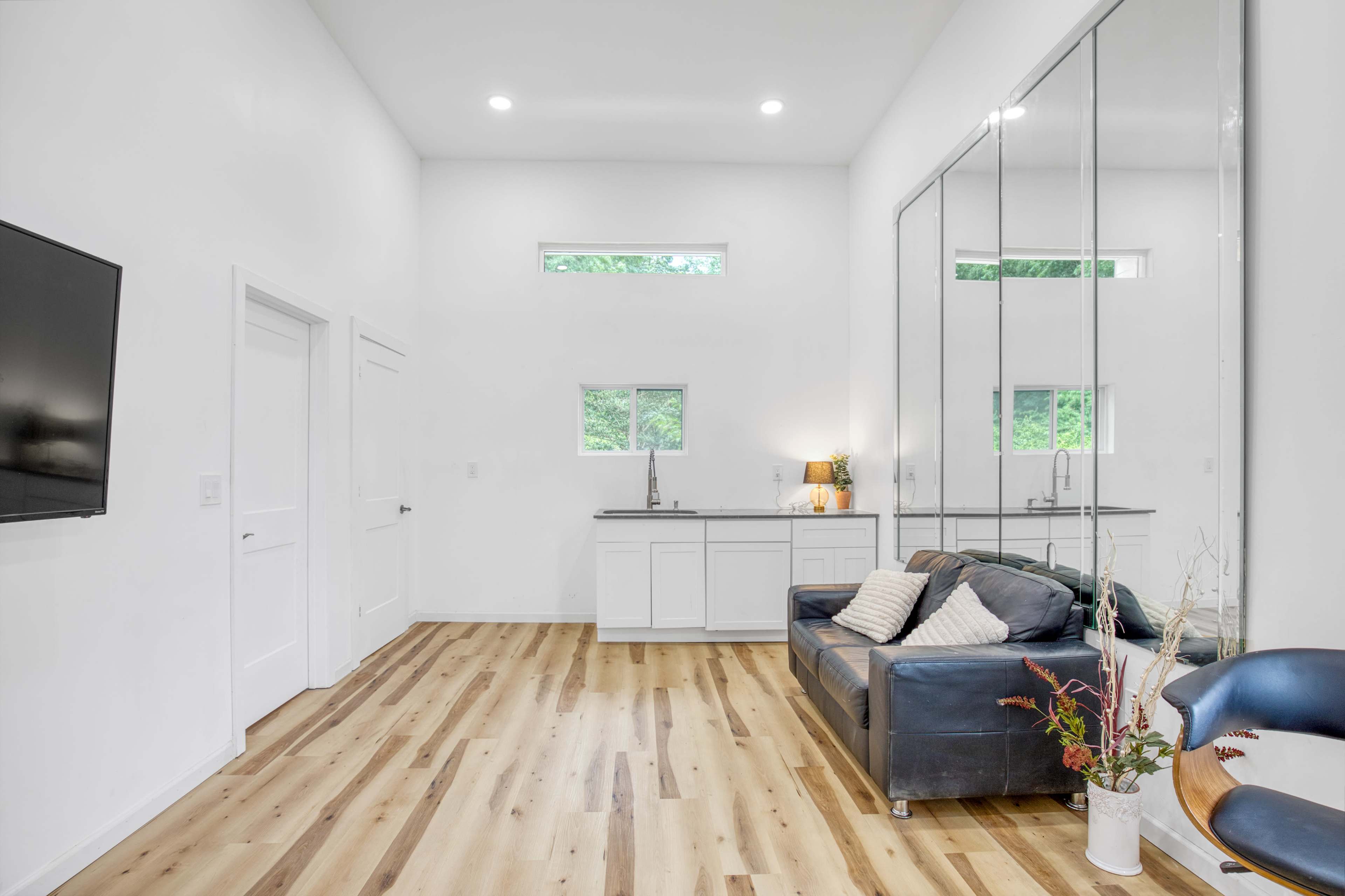 The image shows a modern, spacious interior with light wooden flooring, a black leather couch, a kitchenette area, and large mirrors on one wall.