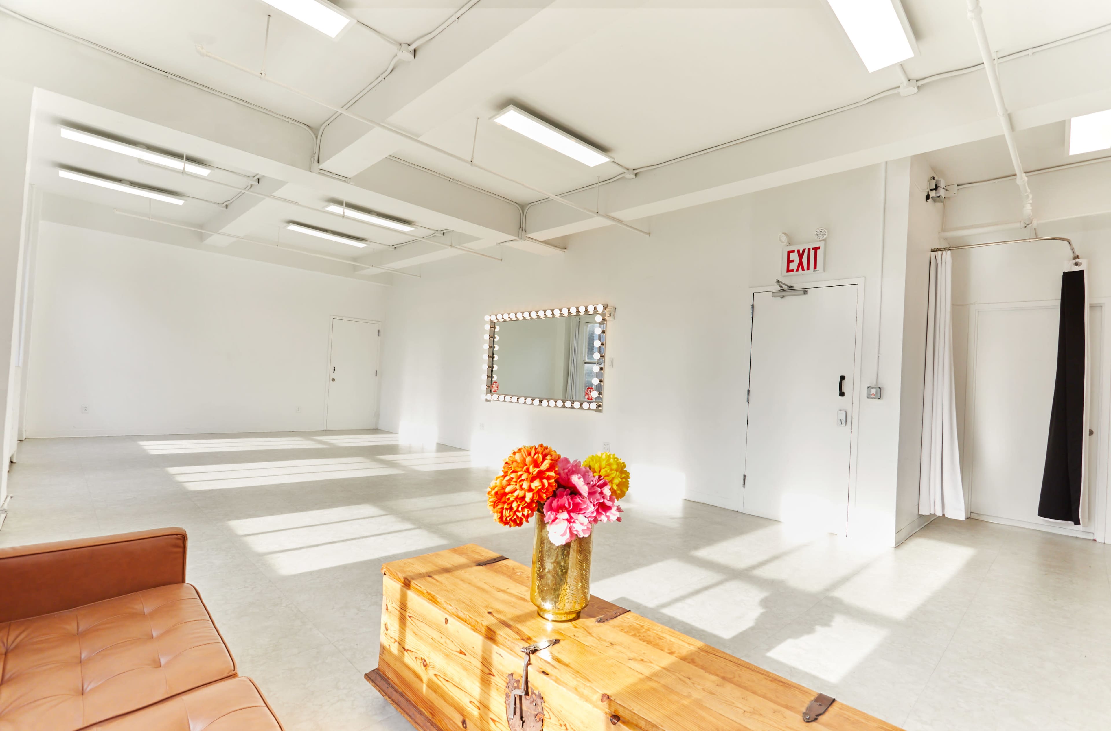 The image shows a spacious, well-lit room with a brown leather couch, a wooden table, and a mirror framed by light bulbs.