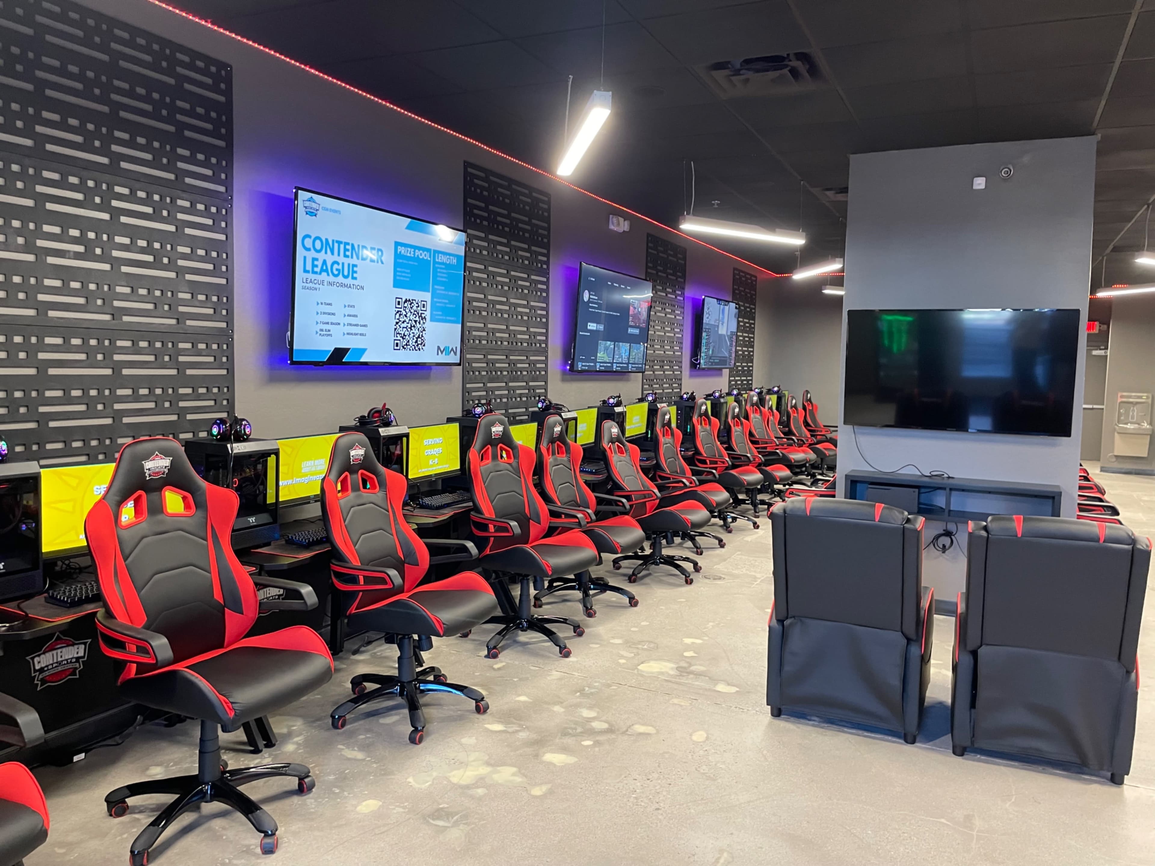 A row of gaming computers with red and black ergonomic chairs is set up in a room featuring multiple large screens on the walls.