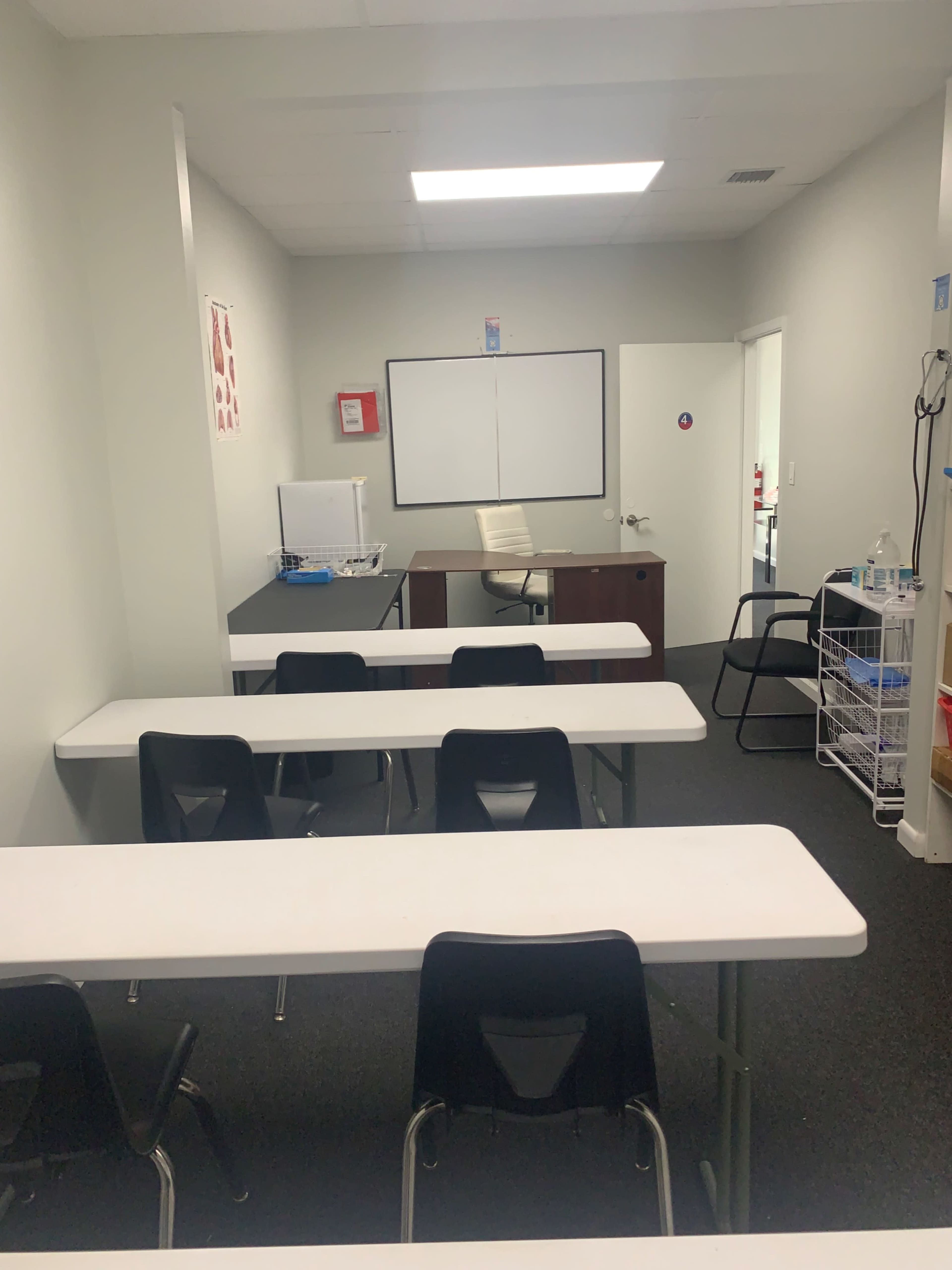A simple classroom with multiple tables and chairs arranged for students, a whiteboard at the front, and a desk along the back wall.