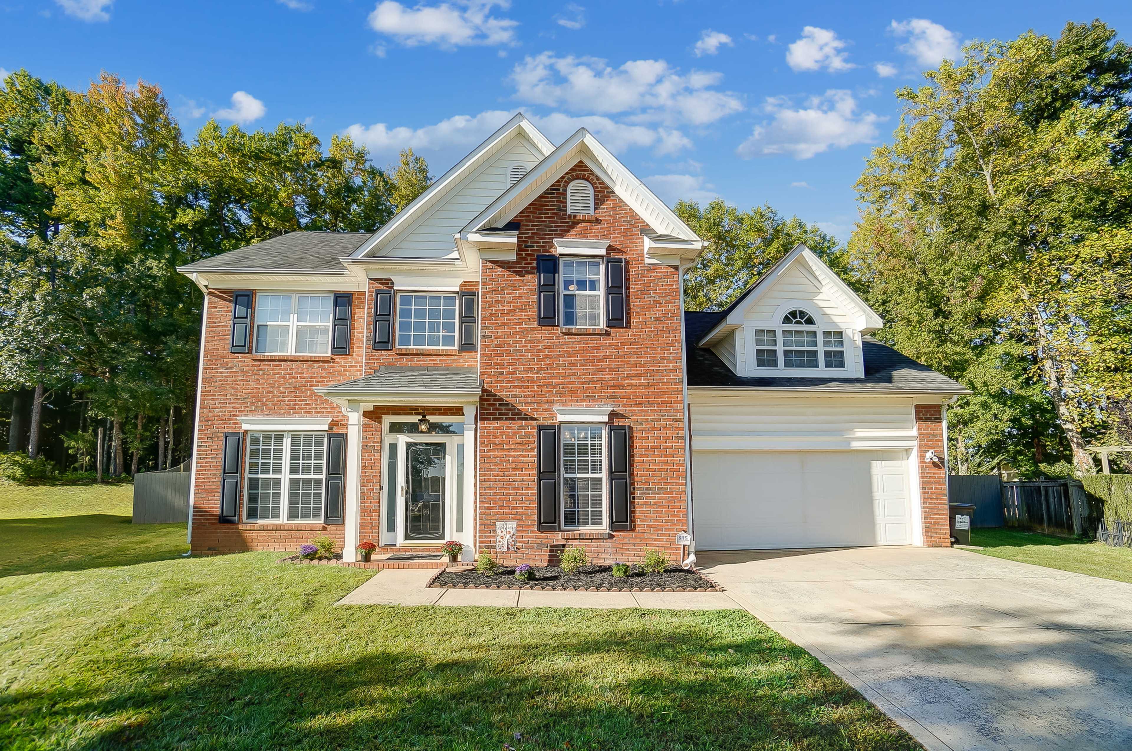 A two-story brick house features a front porch, a two-car garage, and landscaped yard with trees in the background.