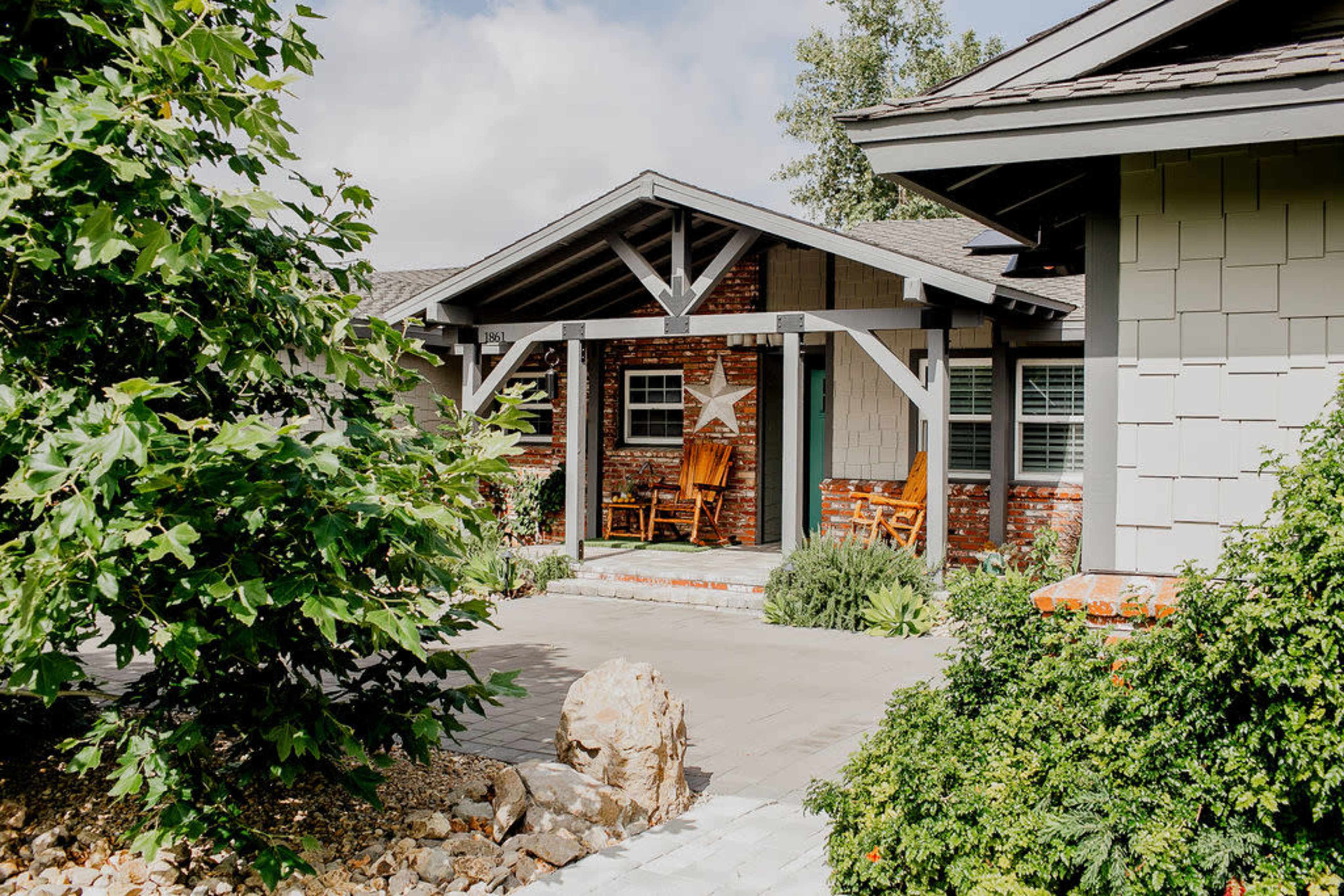 The image shows a front porch of a house with rocking chairs and a star decoration, surrounded by greenery and stone pathways.