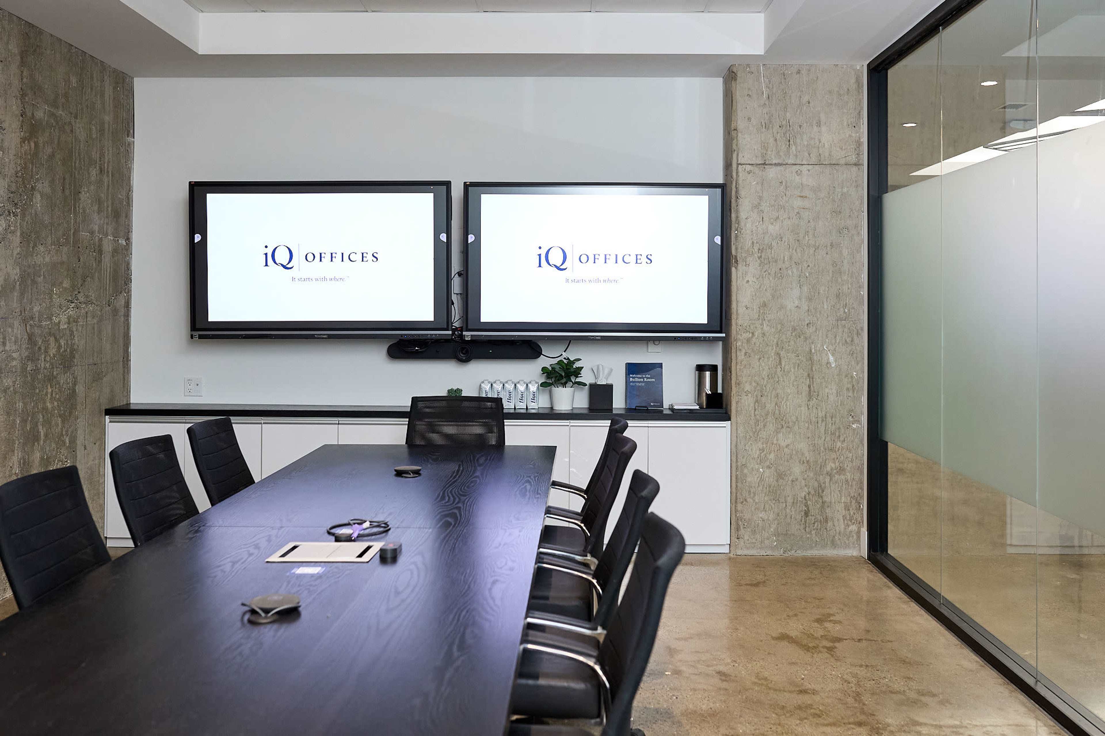 A modern conference room features a long dark table surrounded by black chairs, with two screens displaying the "iQ Offices" logo on the walls.
