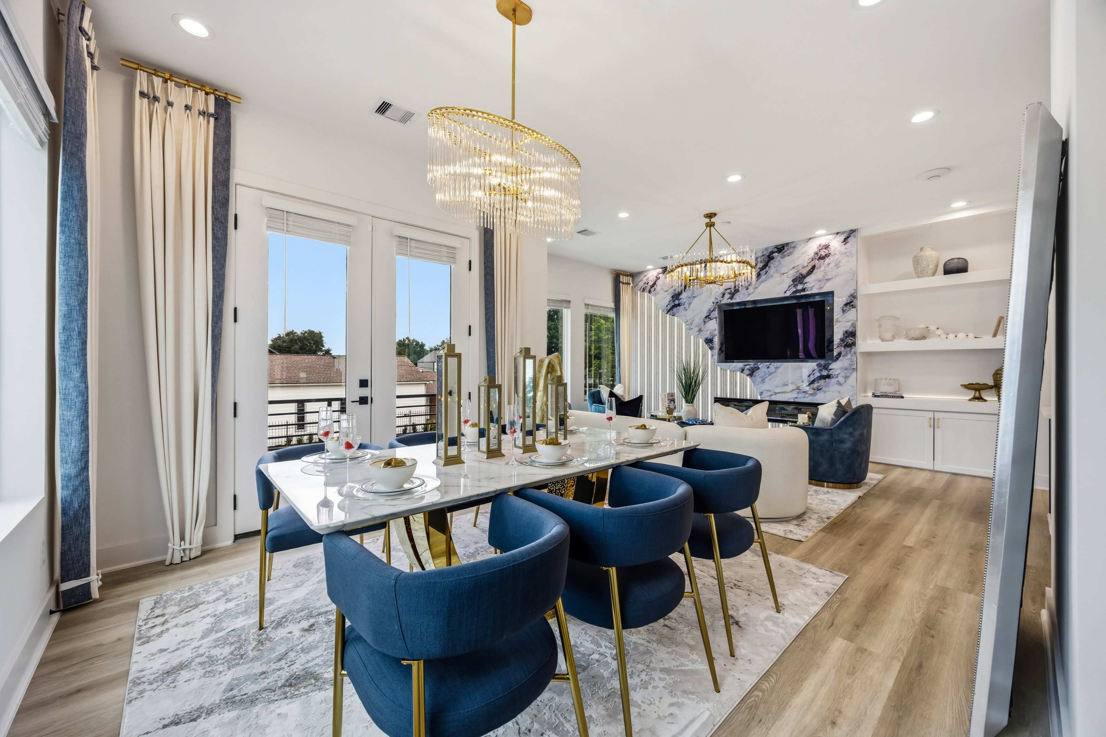 The image shows a modern dining area featuring a large table set for a meal, surrounded by blue chairs, with a contemporary living space visible in the background.