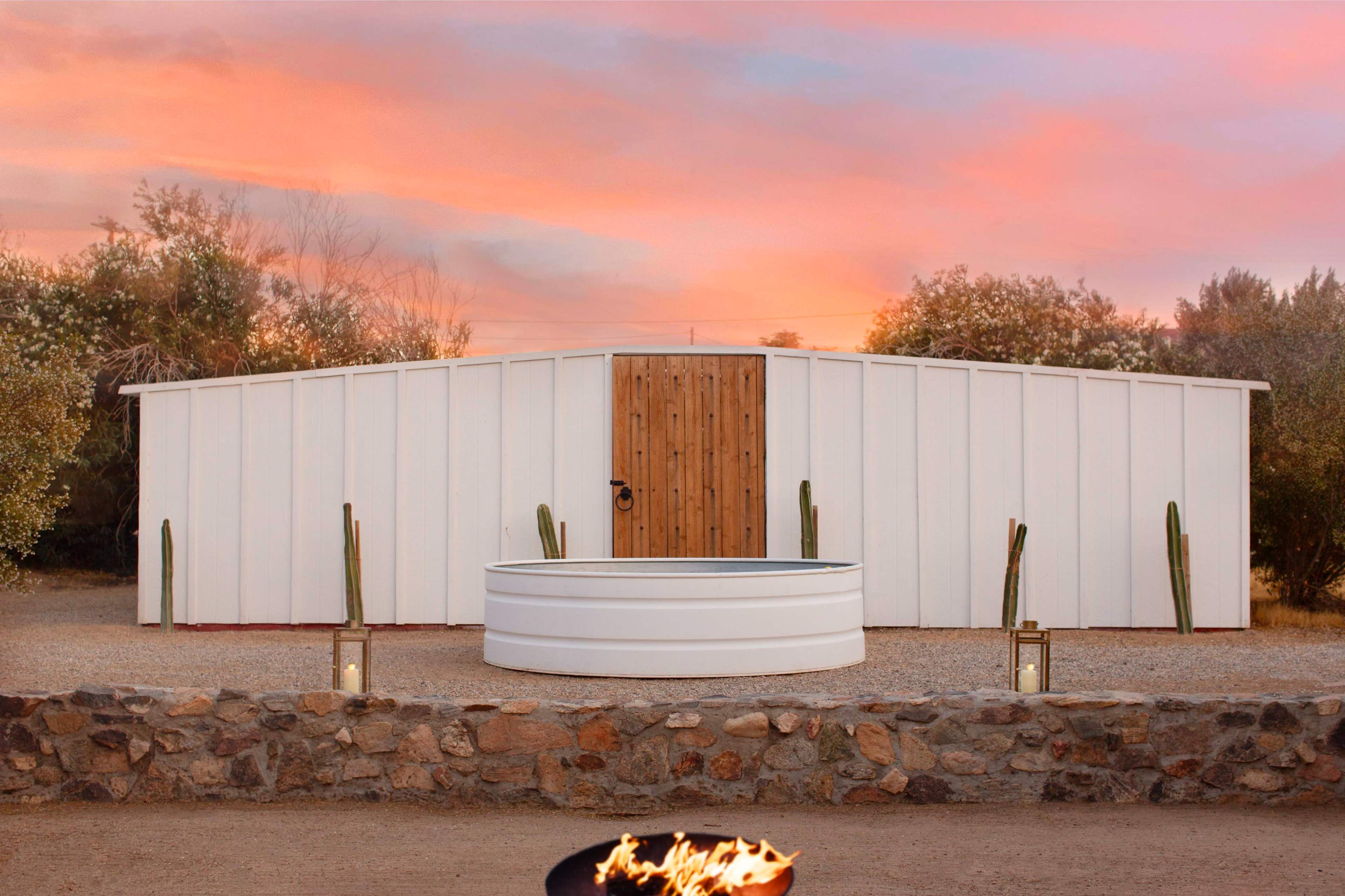 A white modular building with a wooden door is situated in a desert landscape, accompanied by a round, metal tank and cacti in front of a stone wall.