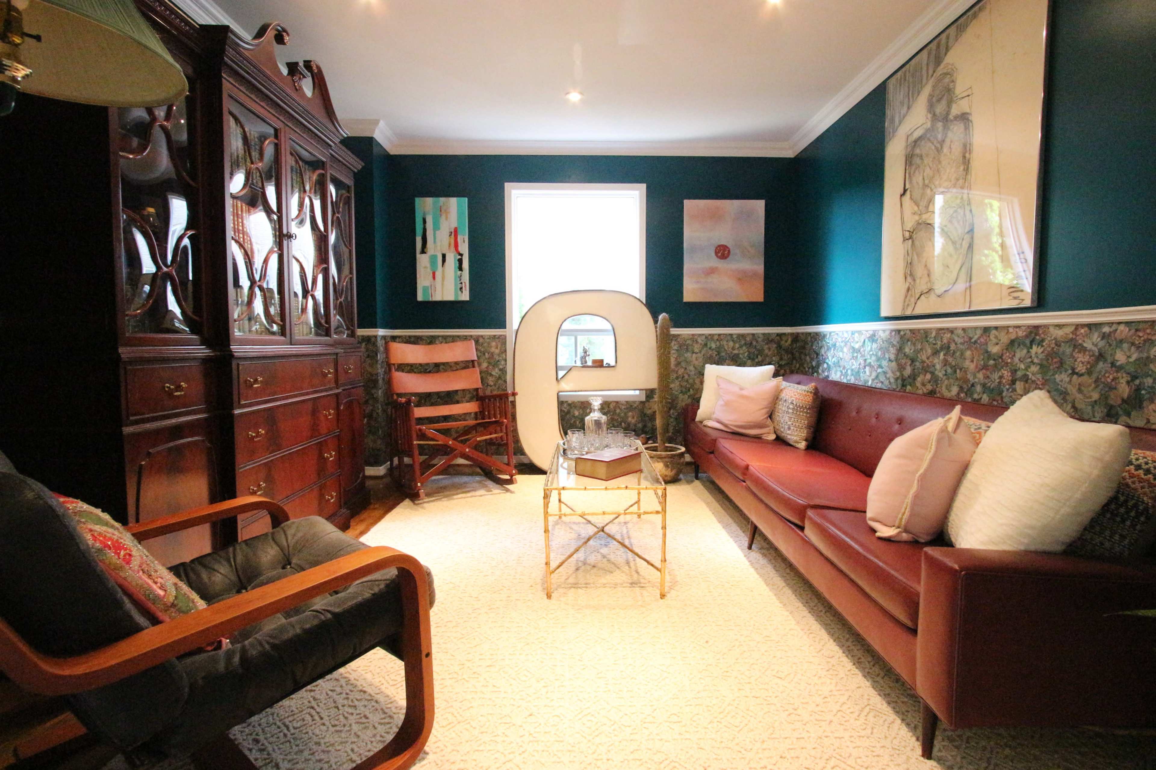 The image shows a cozy living room featuring a dark wooden cabinet, a leather sofa, a chair, and a coffee table, with floral wallpaper and artwork on the walls.