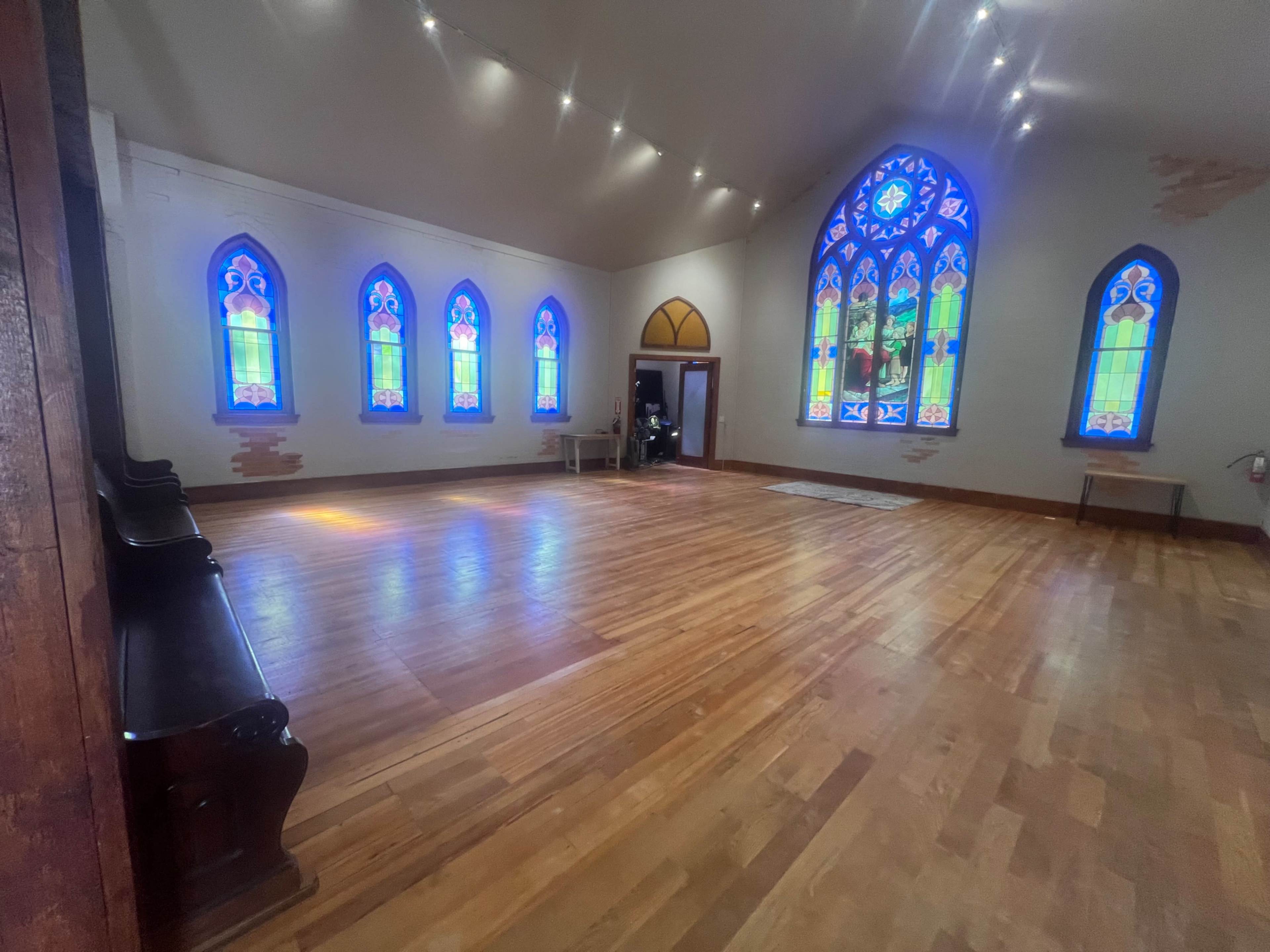 The image shows an empty room with wooden flooring and colorful stained glass windows along the walls.