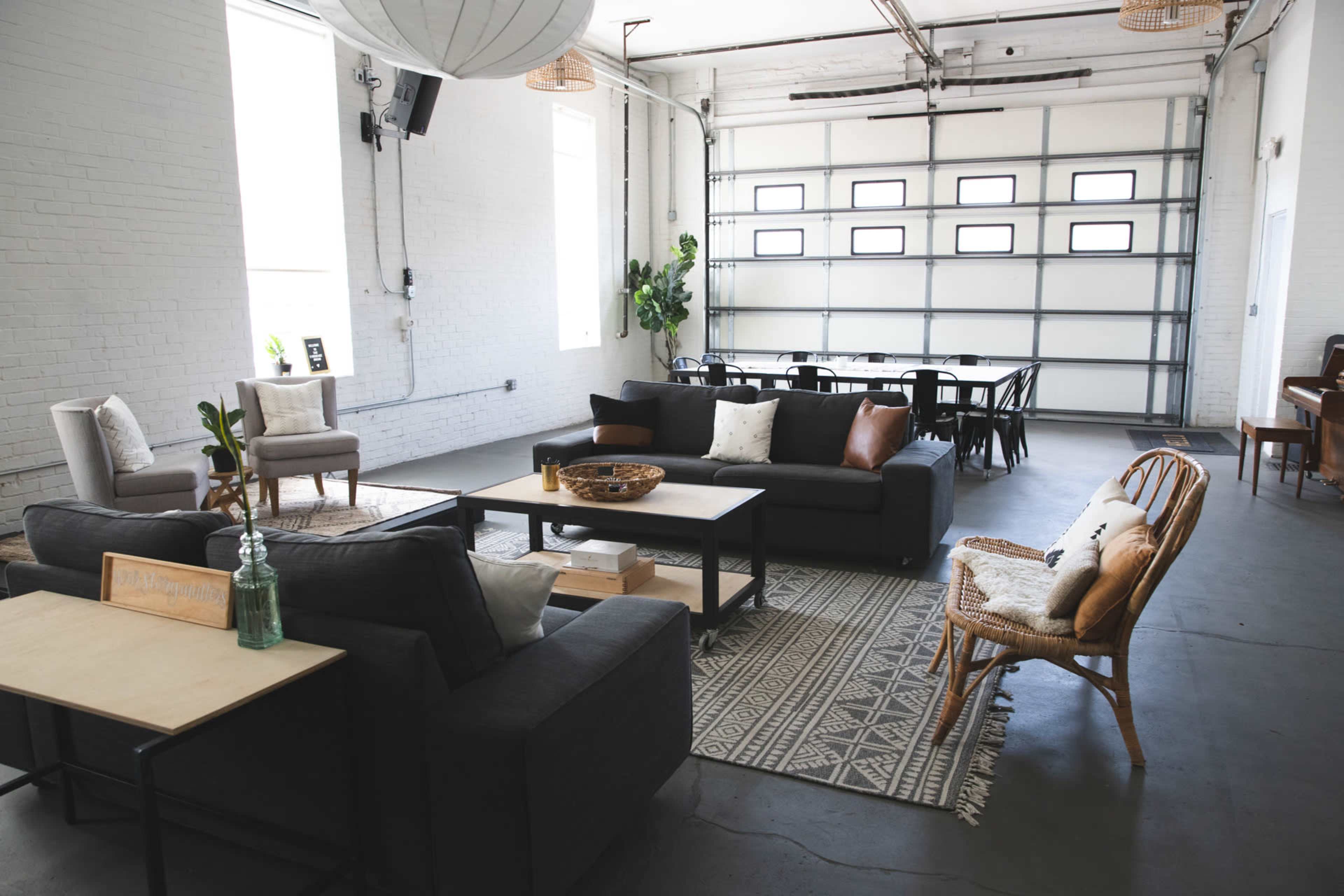 A spacious indoor area with several seating arrangements, including gray sofas and a wooden table, surrounded by large windows and a garage door.