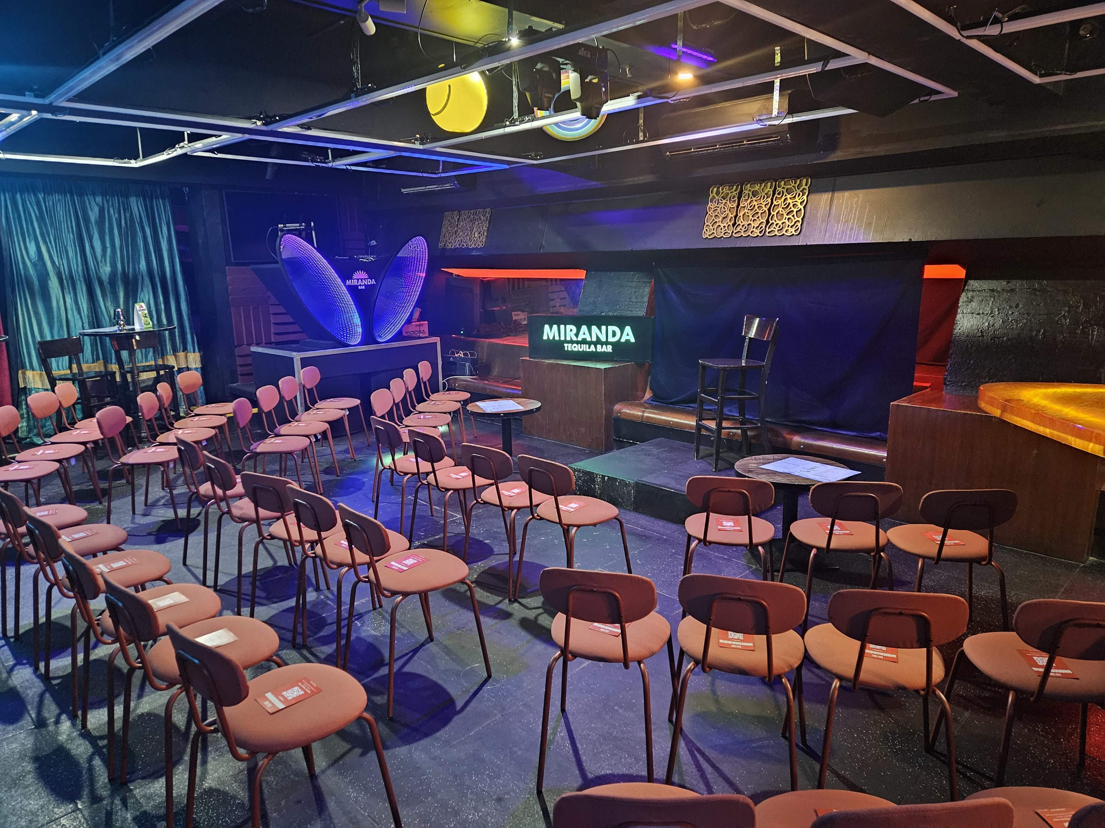 The image shows a small performance venue with rows of empty chairs facing a stage that has a sign reading "MIRANDA" and a stool positioned on it.