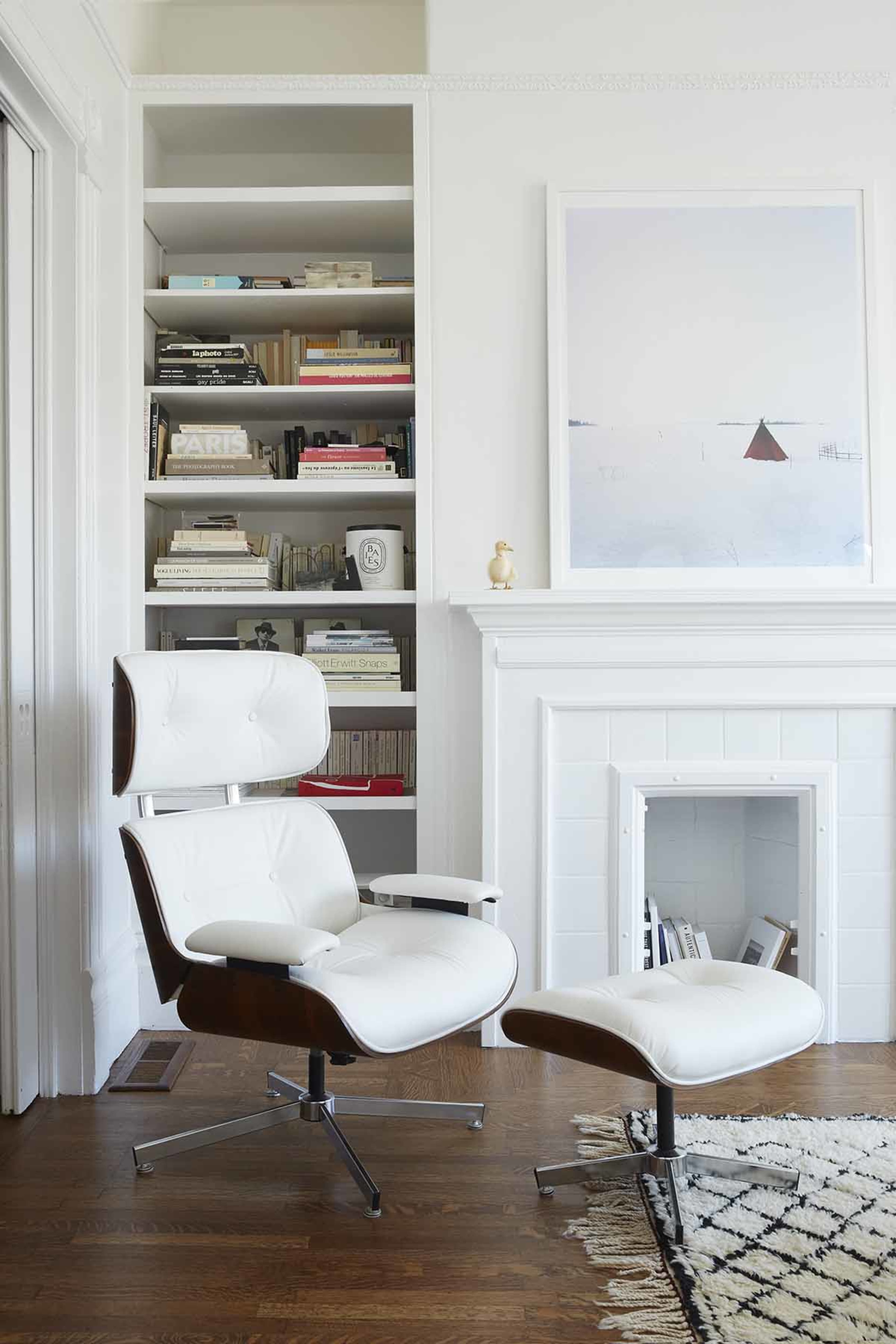 A white lounge chair with an ottoman is positioned next to a built-in bookshelf filled with books, with a minimalist artwork displayed on the wall above a white fireplace.