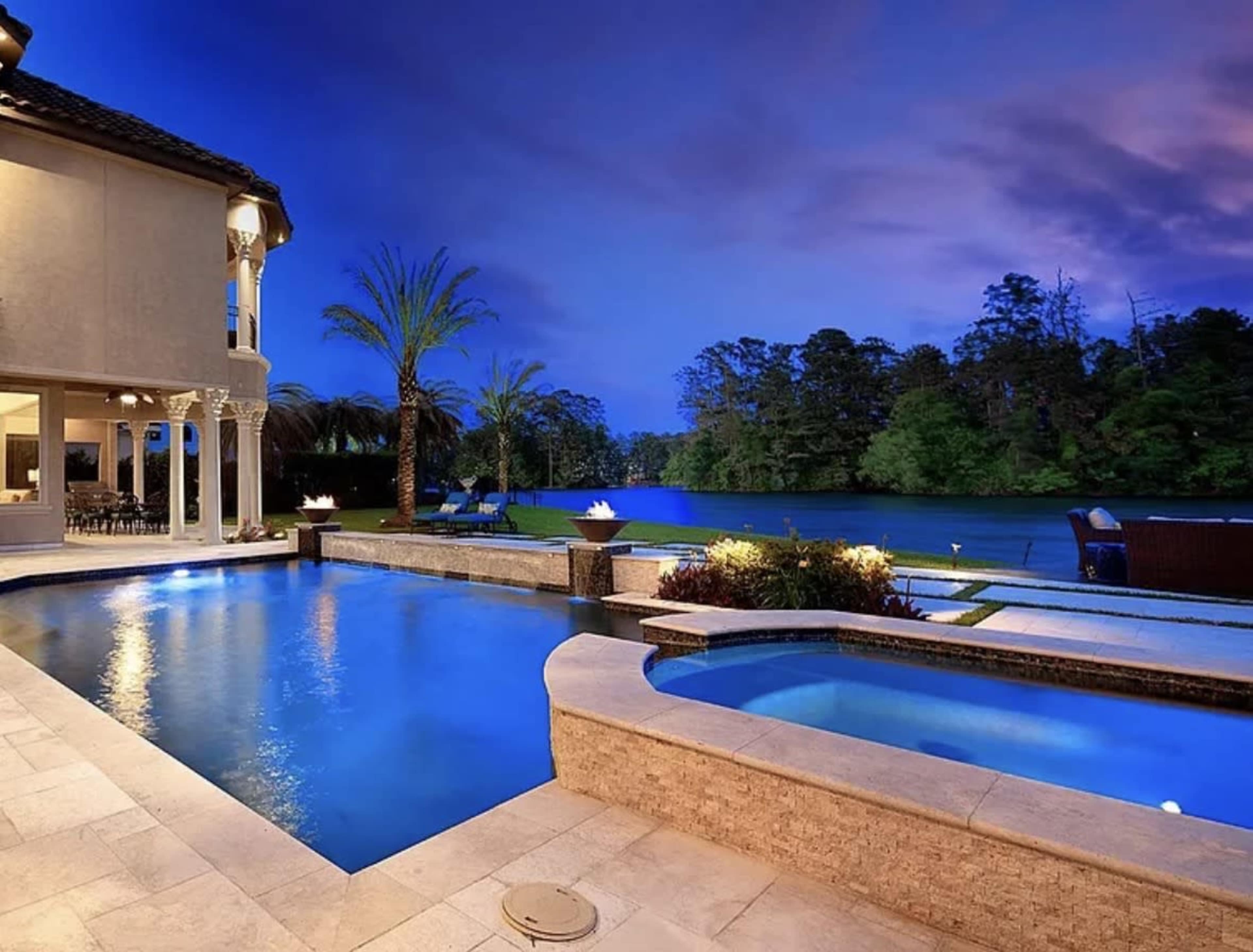 A well-lit backyard features a pool and spa overlooking a lake, surrounded by palm trees and lush greenery under a twilight sky.