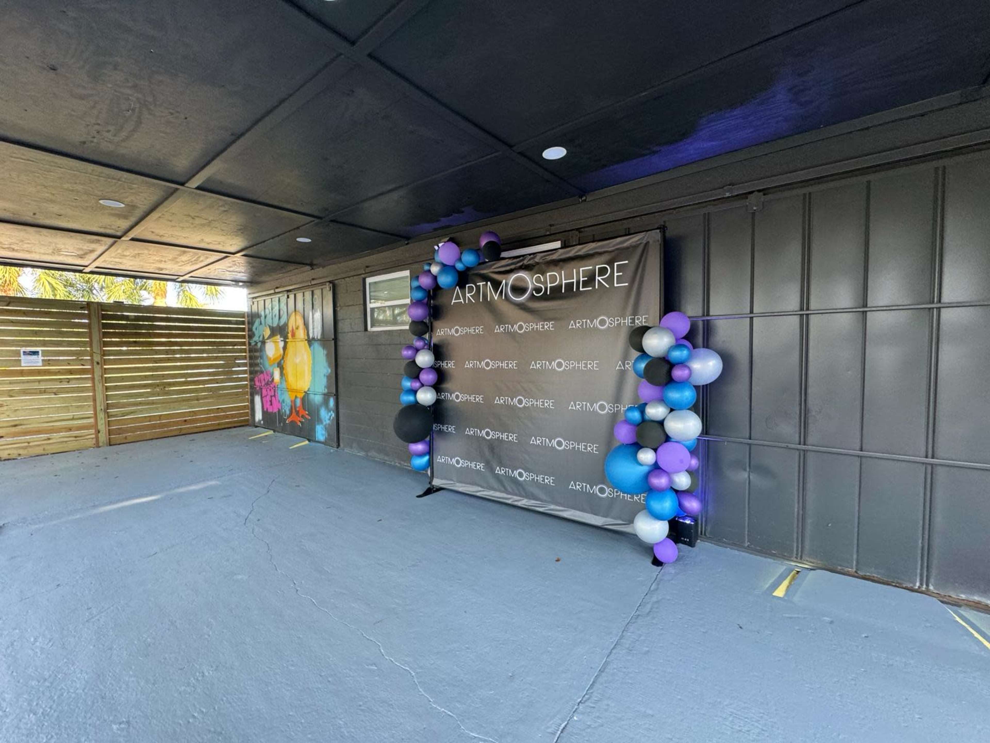 A decorated event space featuring a backdrop with the word "ARTMOSPHERE" and a cluster of purple, white, and blue balloons.