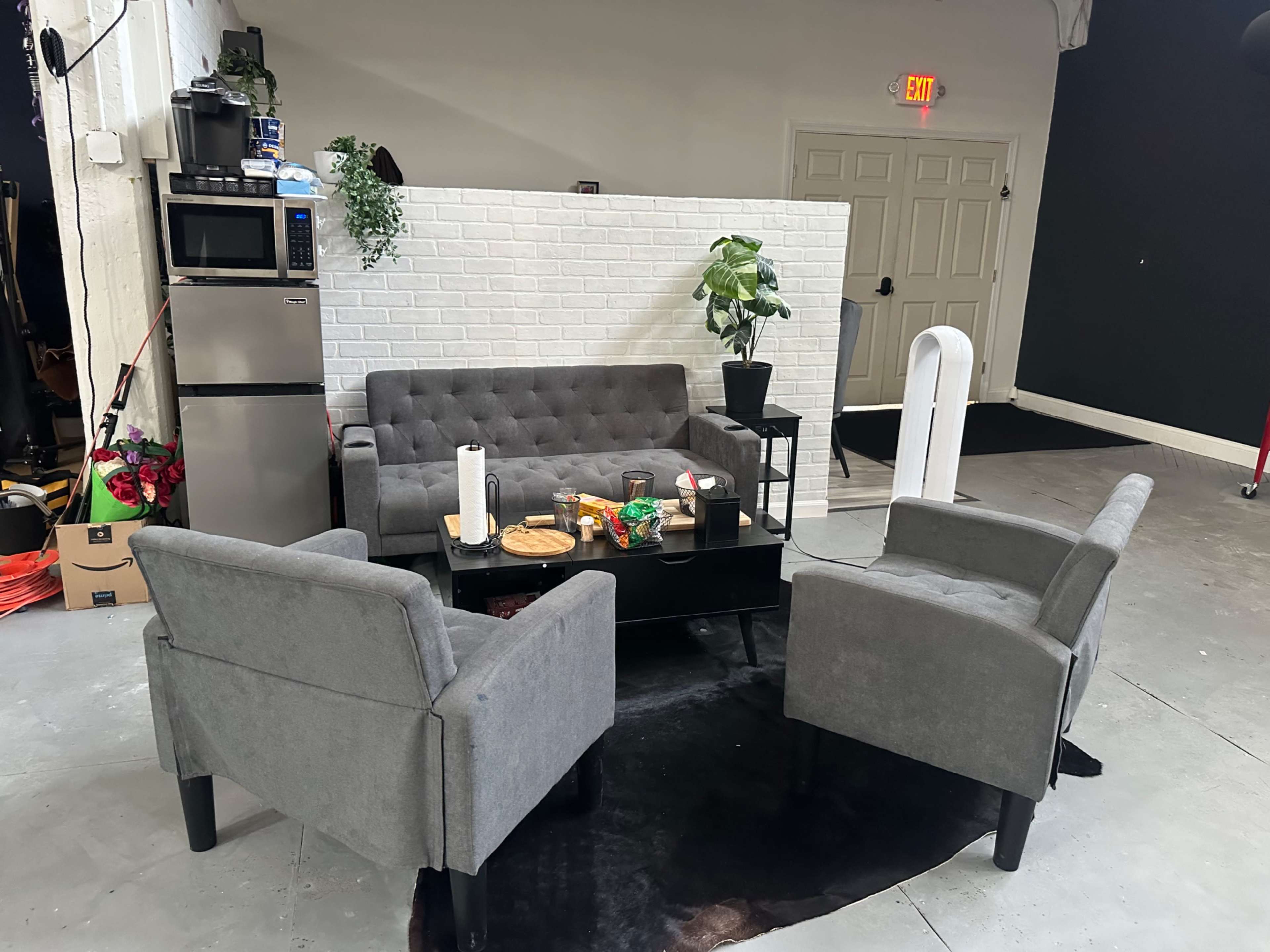 The image shows a cozy seating area with a gray sofa and two armchairs arranged around a black coffee table in front of a white brick wall.
