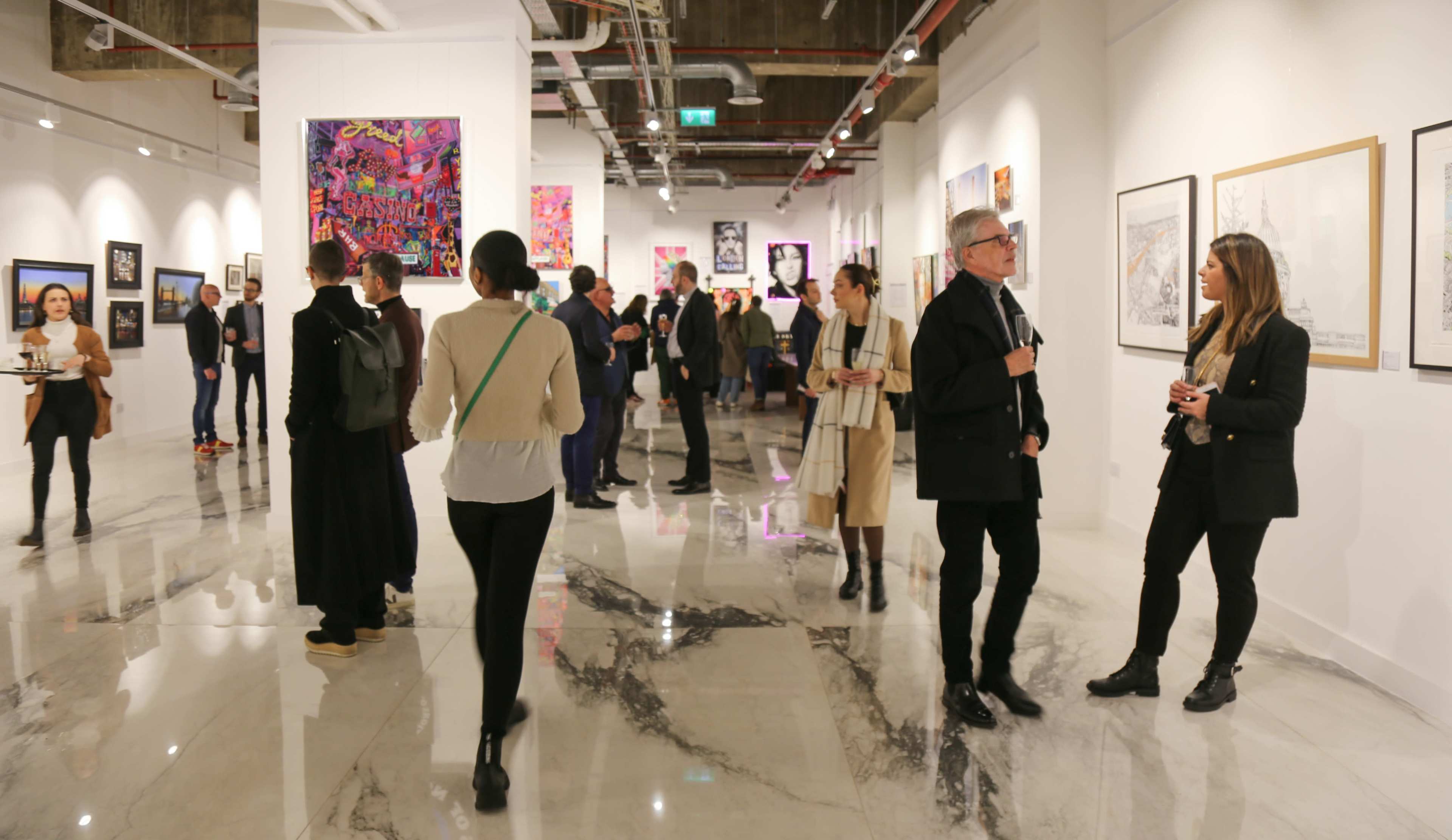 A group of people mingles in an art gallery while observing various colorful artworks displayed on the walls.