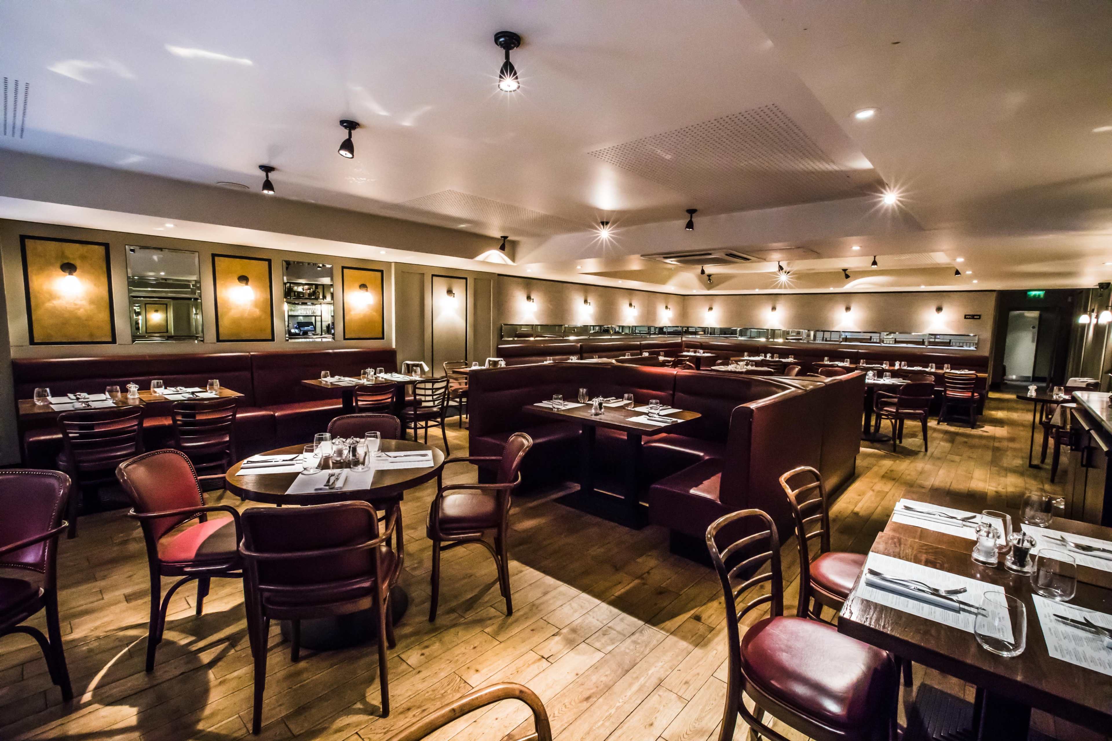 The image shows a spacious restaurant with wooden floors, wooden tables, and red upholstered seating arranged around various tables and booths.