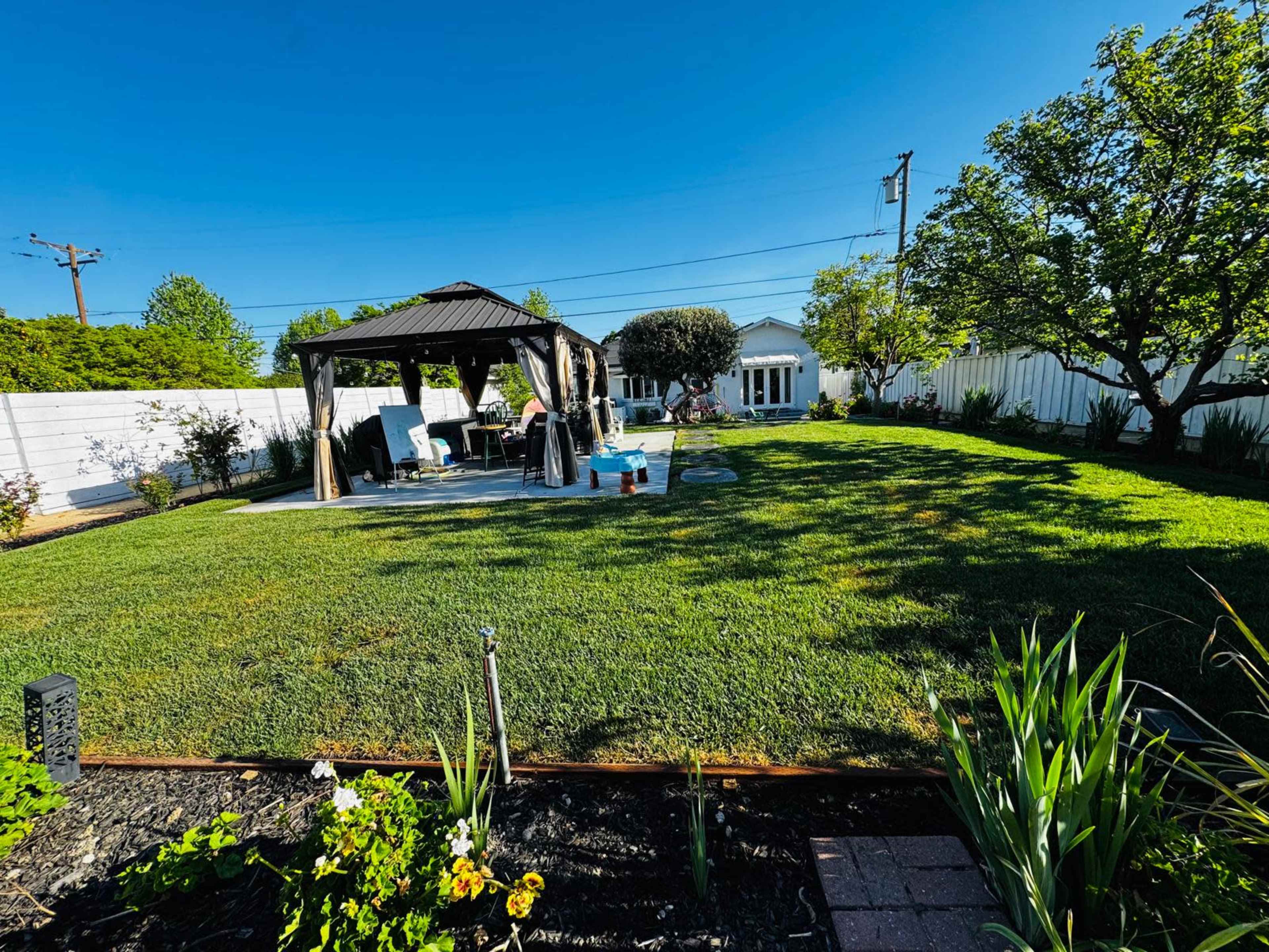 The image shows a spacious backyard featuring a gazebo, seating area, a well-maintained lawn, and a white fence surrounding the property.