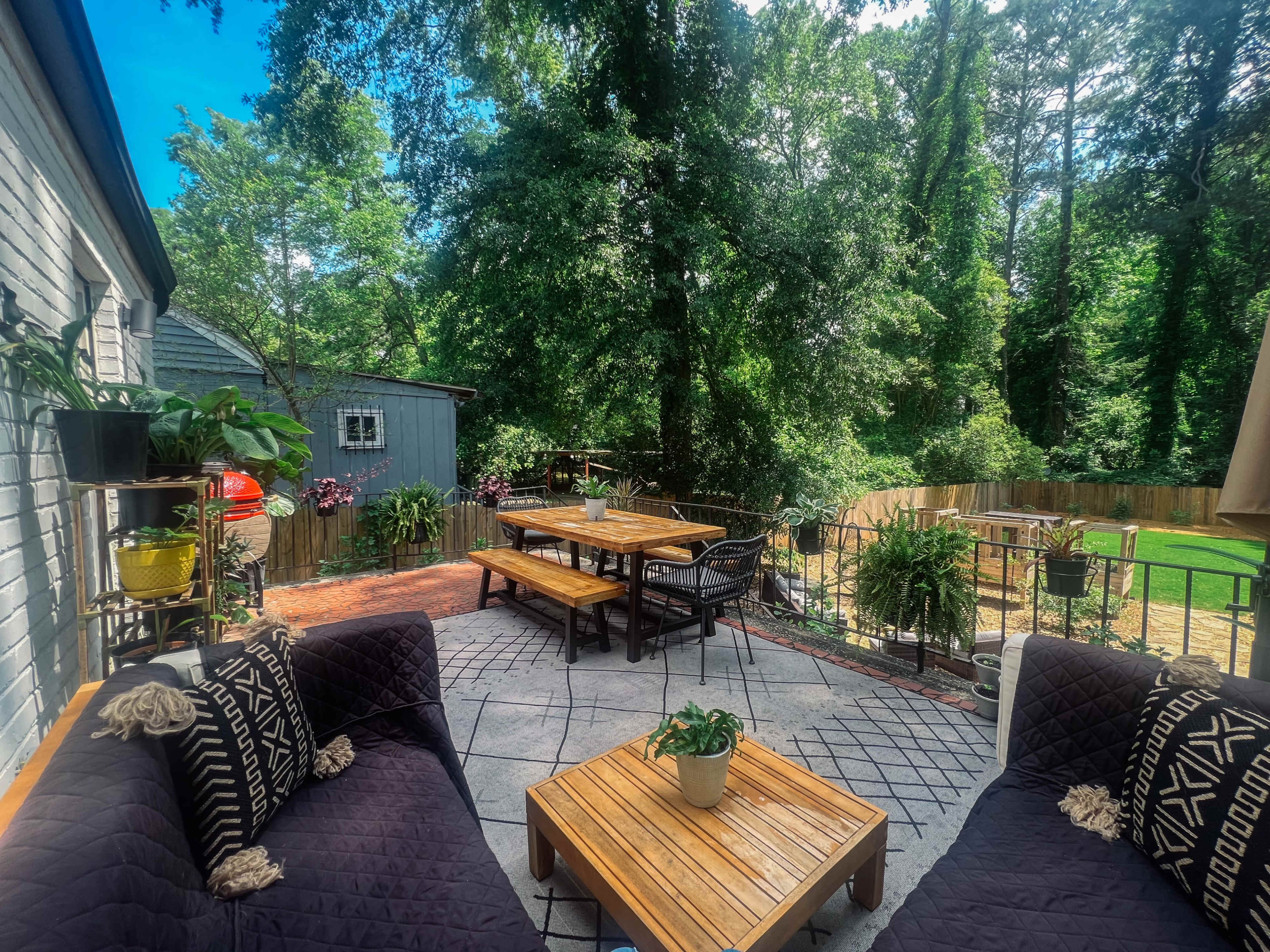 A patio area features wooden furniture surrounded by greenery and a fence in the background.