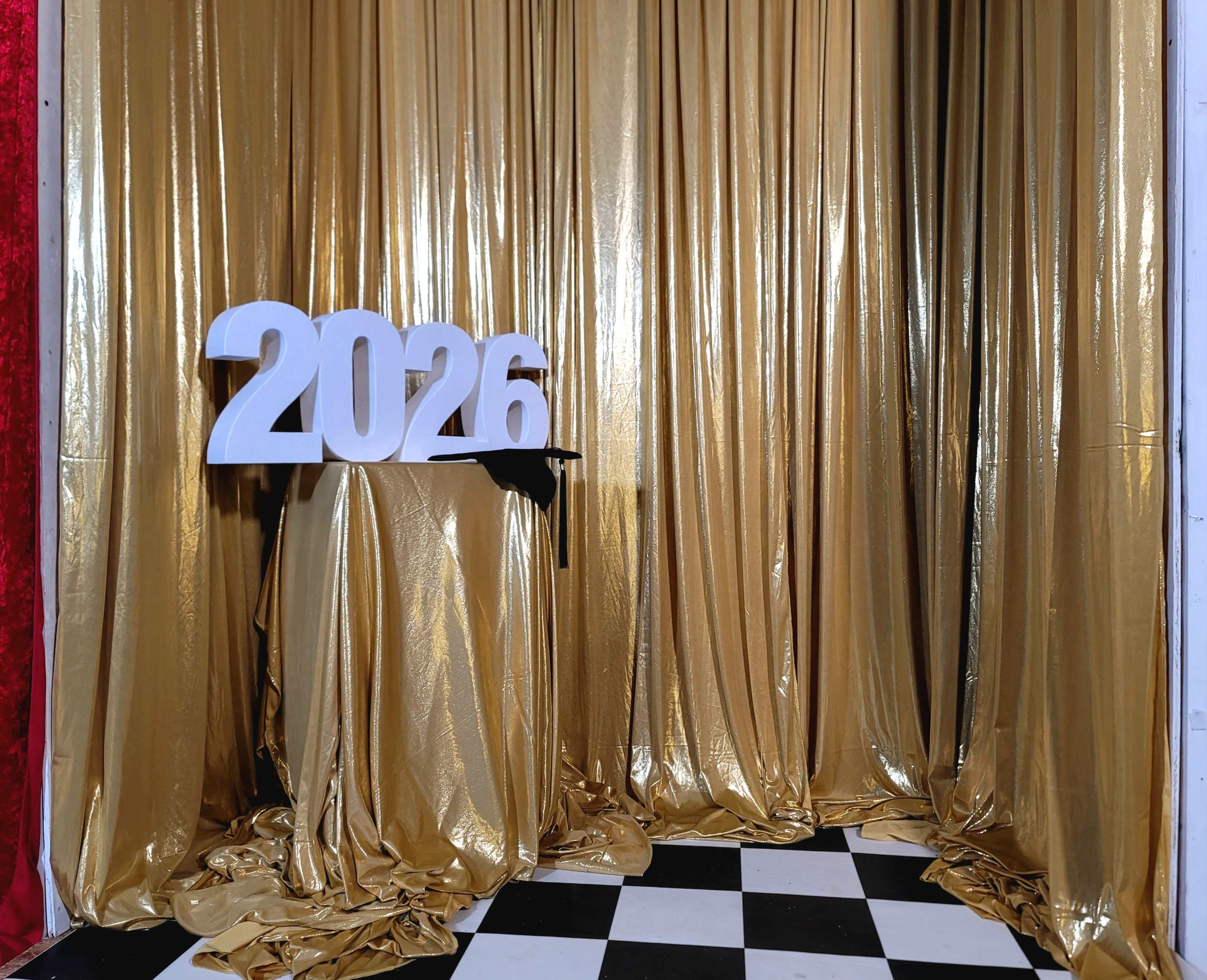 The image shows a gold curtain backdrop with the numbers "2026" displayed on a pedestal in front of a checkered floor.