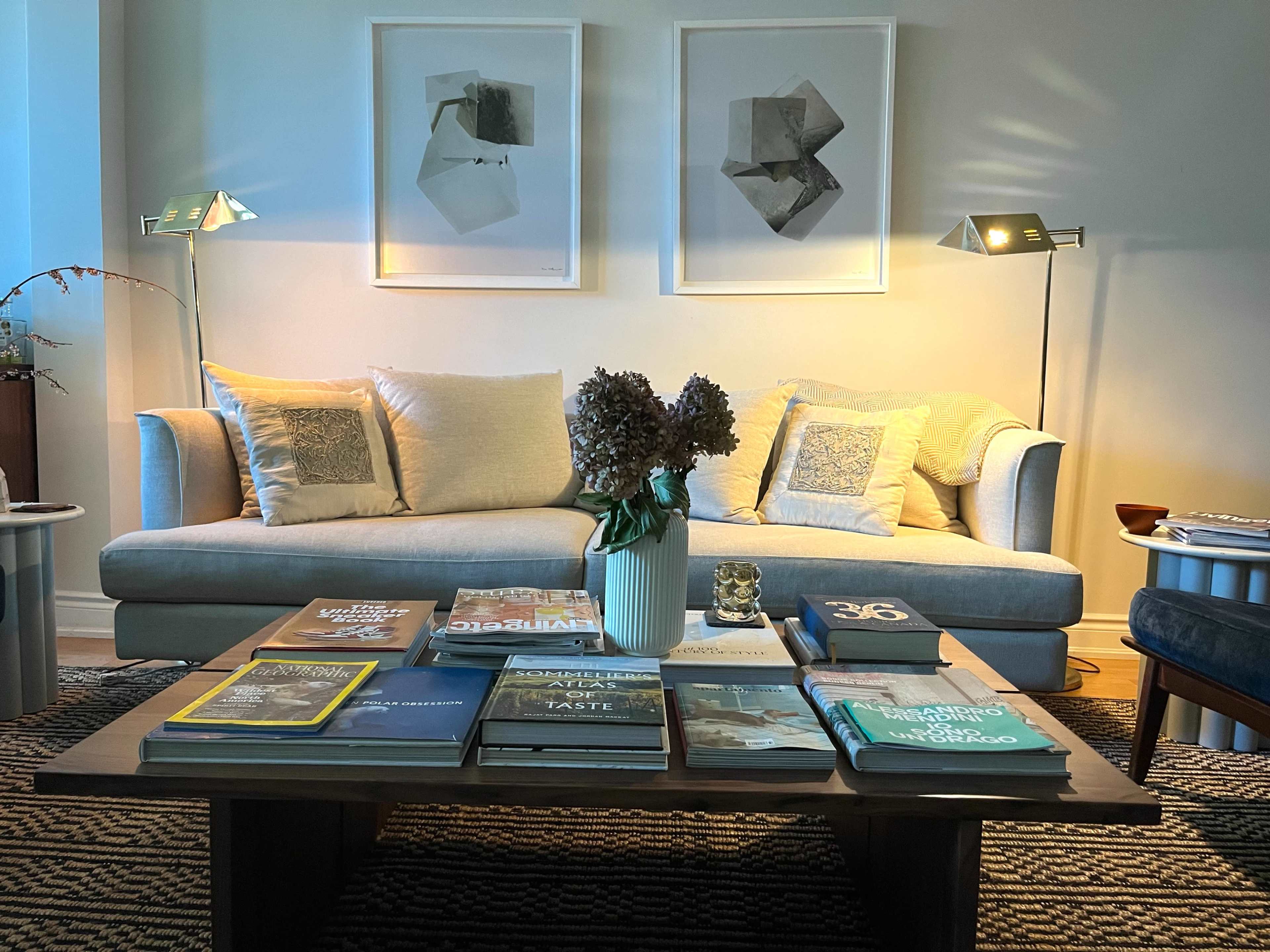 A modern living room features a light-colored sofa with decorative pillows, a wooden coffee table stacked with books, and two framed artworks on the wall, illuminated by stylish floor lamps.