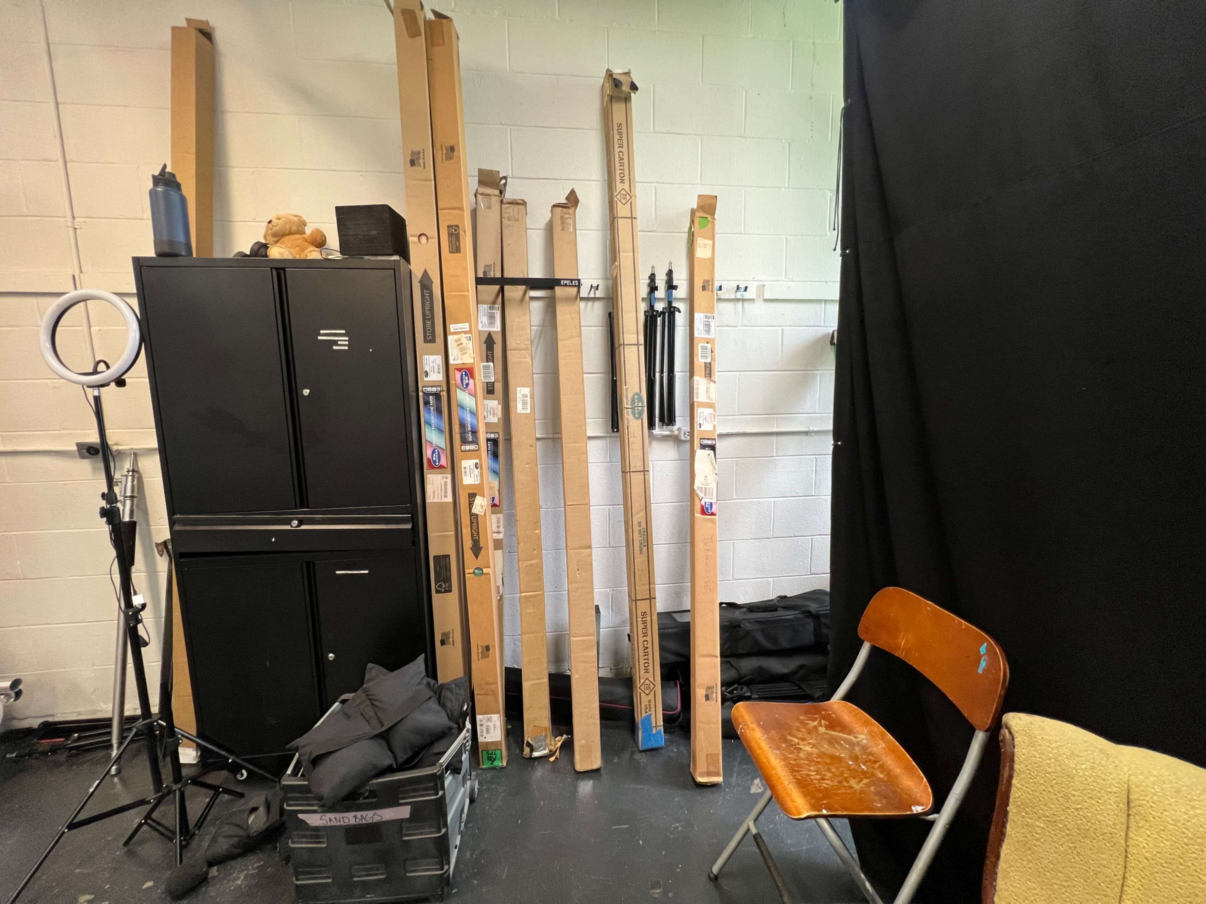 The image shows cardboard tubes leaning against a black storage cabinet in a studio space, with a folding chair and other items on the floor.