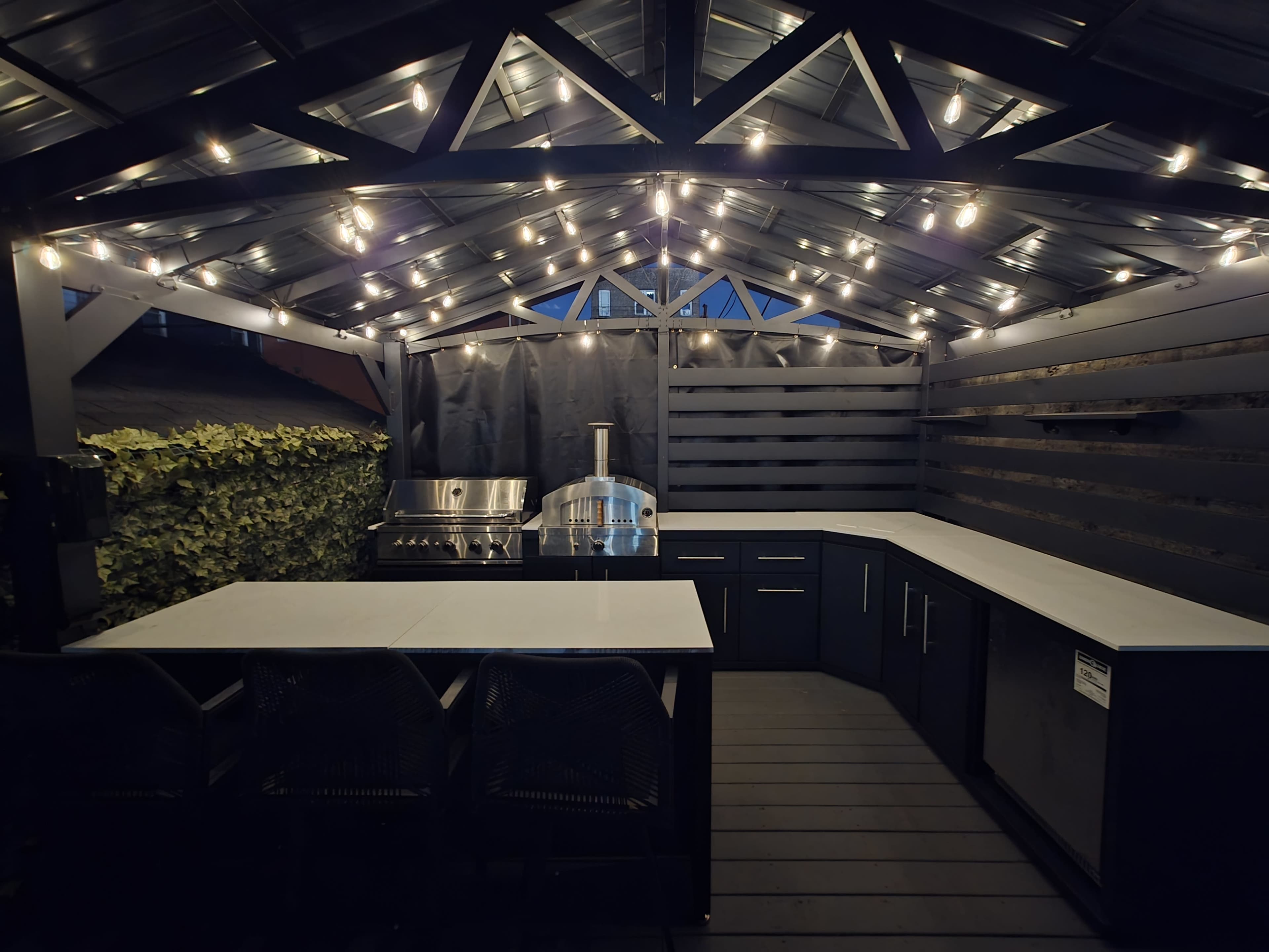 The image shows a modern outdoor kitchen under a covered structure with string lights, featuring a grill, a pizza oven, and a kitchen island with seating.