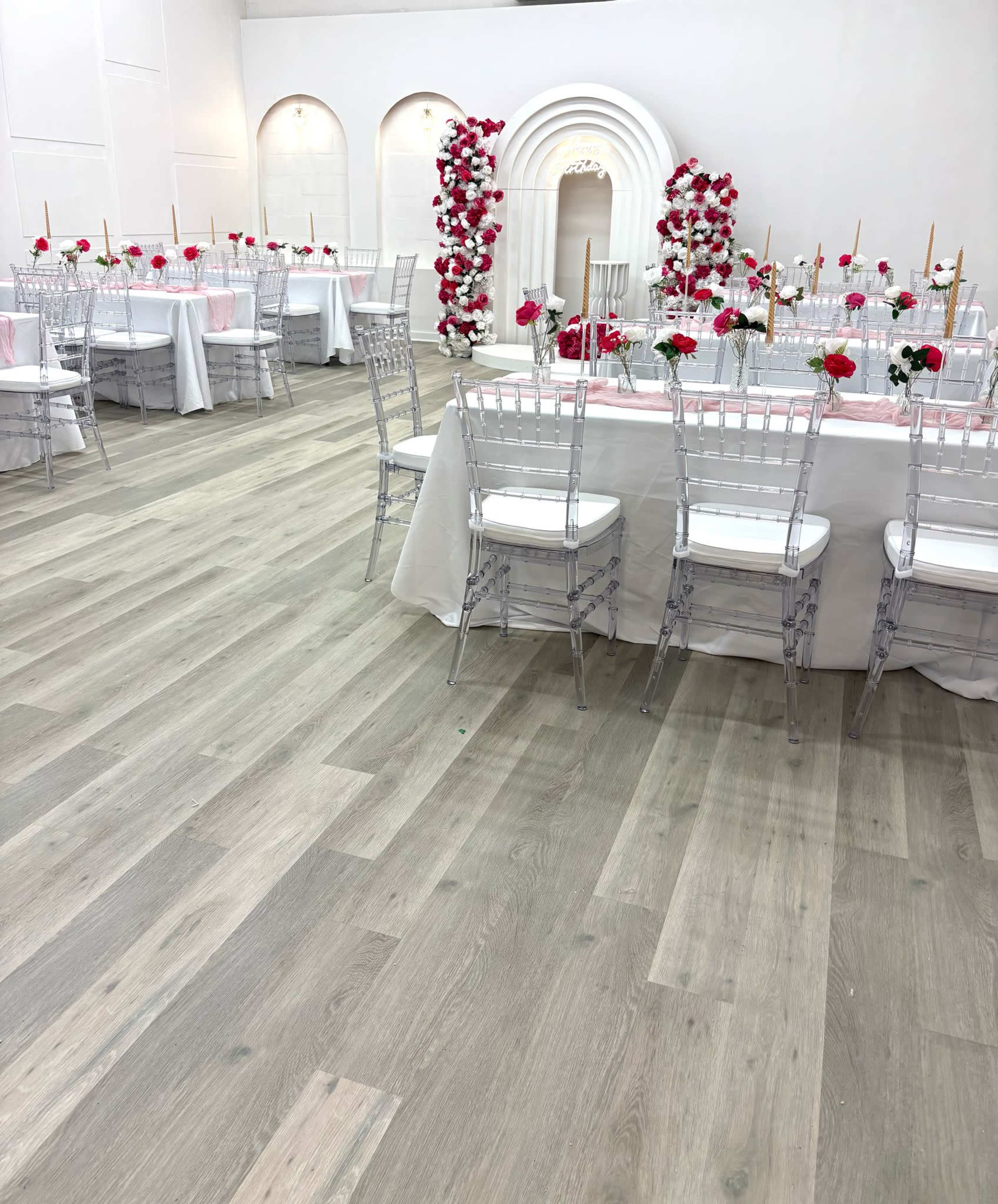 The image shows a spacious event venue set up for a celebration, featuring white tables adorned with floral centerpieces and transparent chairs.