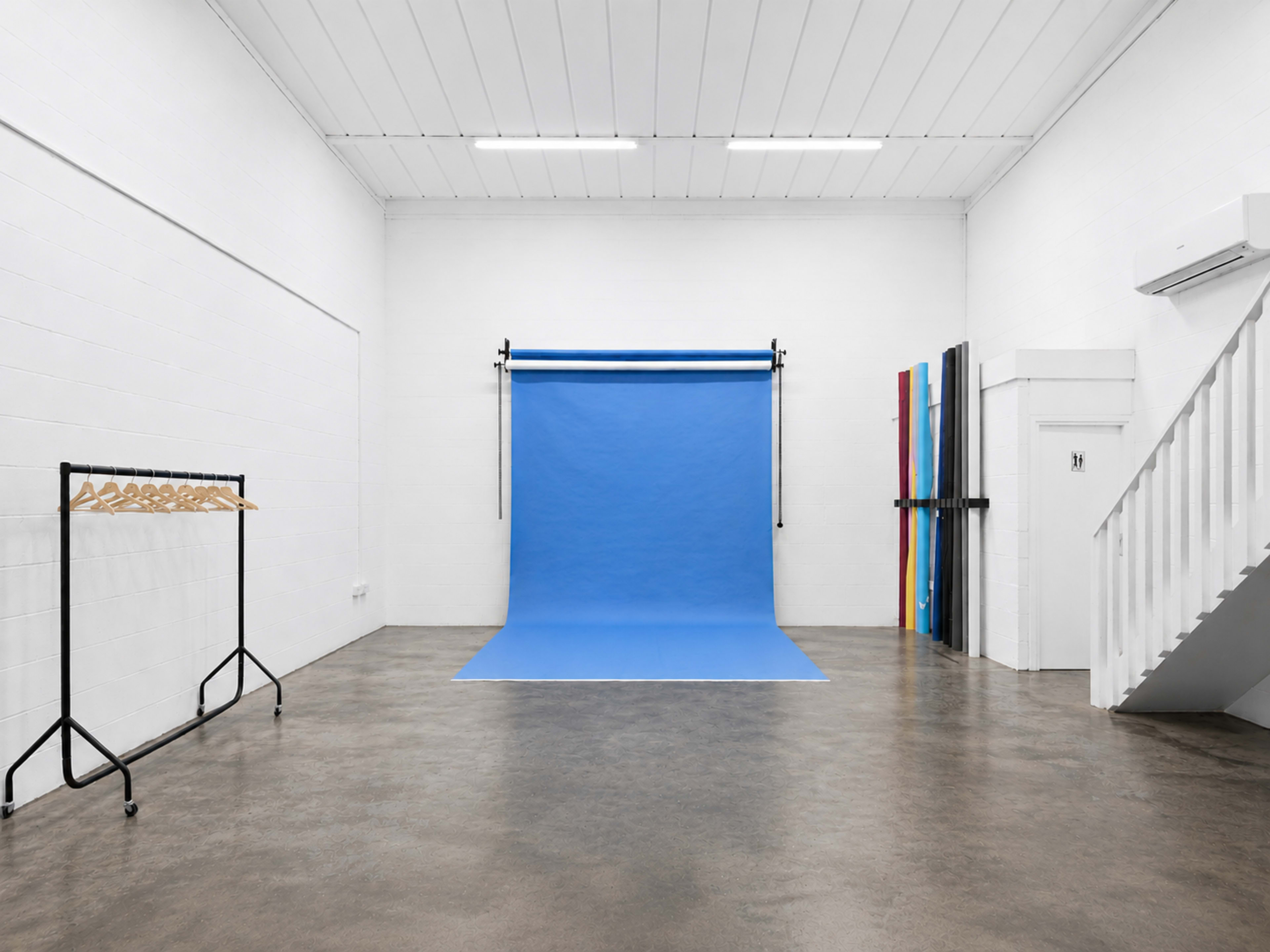The image shows a spacious, brightly lit studio featuring a blue backdrop, a clothing rack on the left, and colorful panels on the right near a staircase.