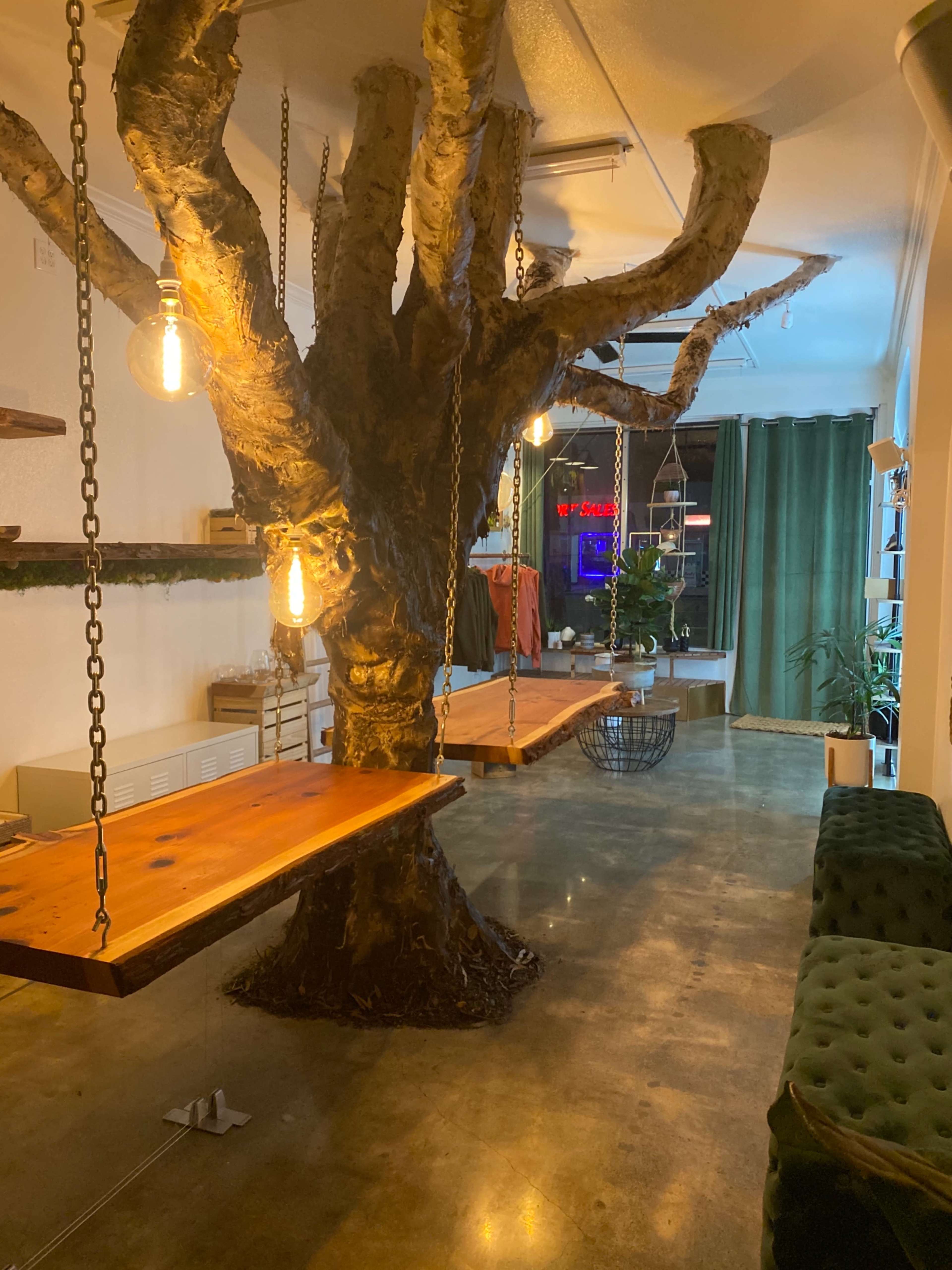 A room with a large, indoor tree sculpture at its center, from which wooden swings are suspended, surrounded by green curtains and various plants.