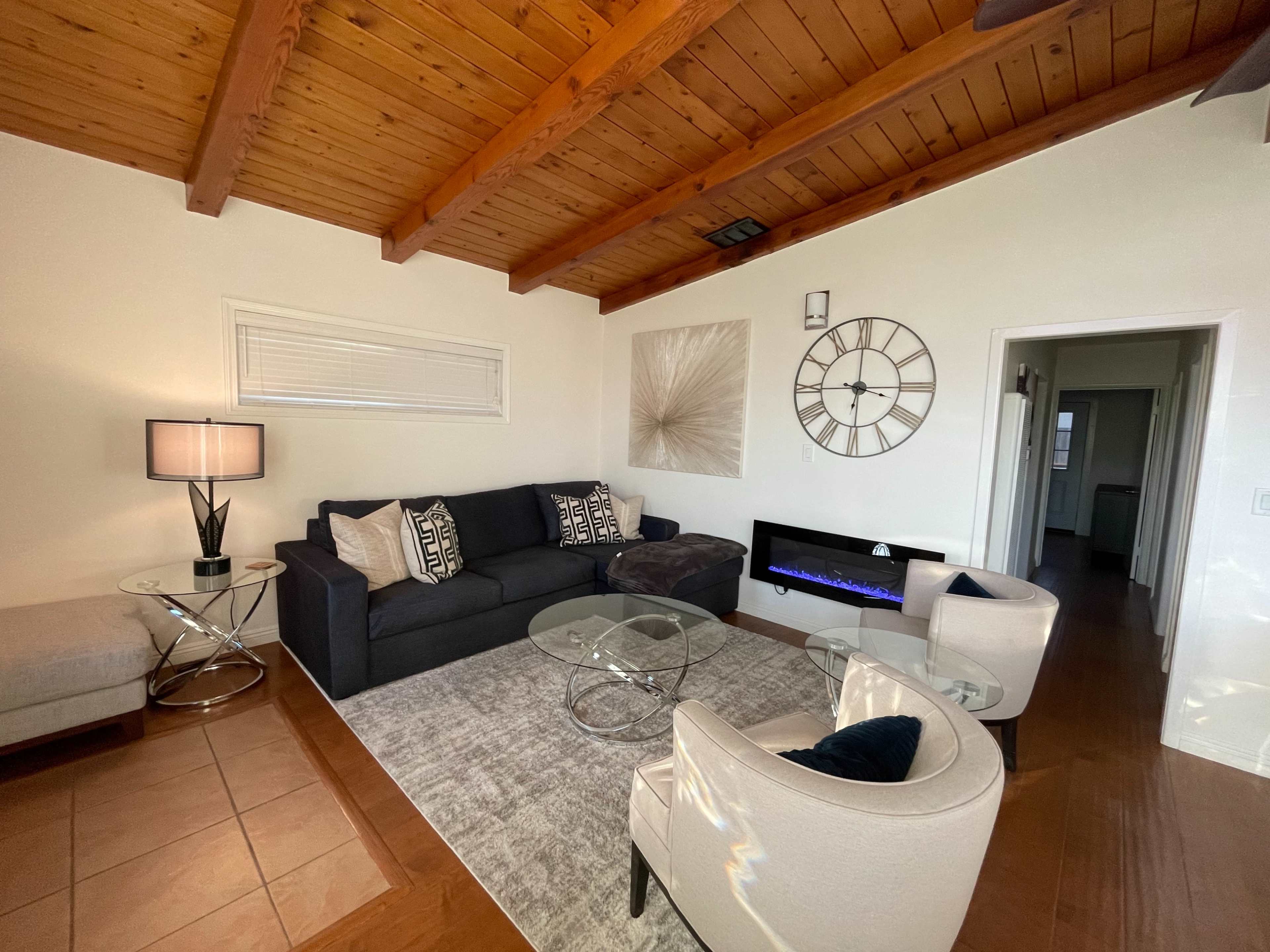 A living room with a dark sofa, a glass coffee table, two armchairs, and a modern electric fireplace beneath a wall clock.
