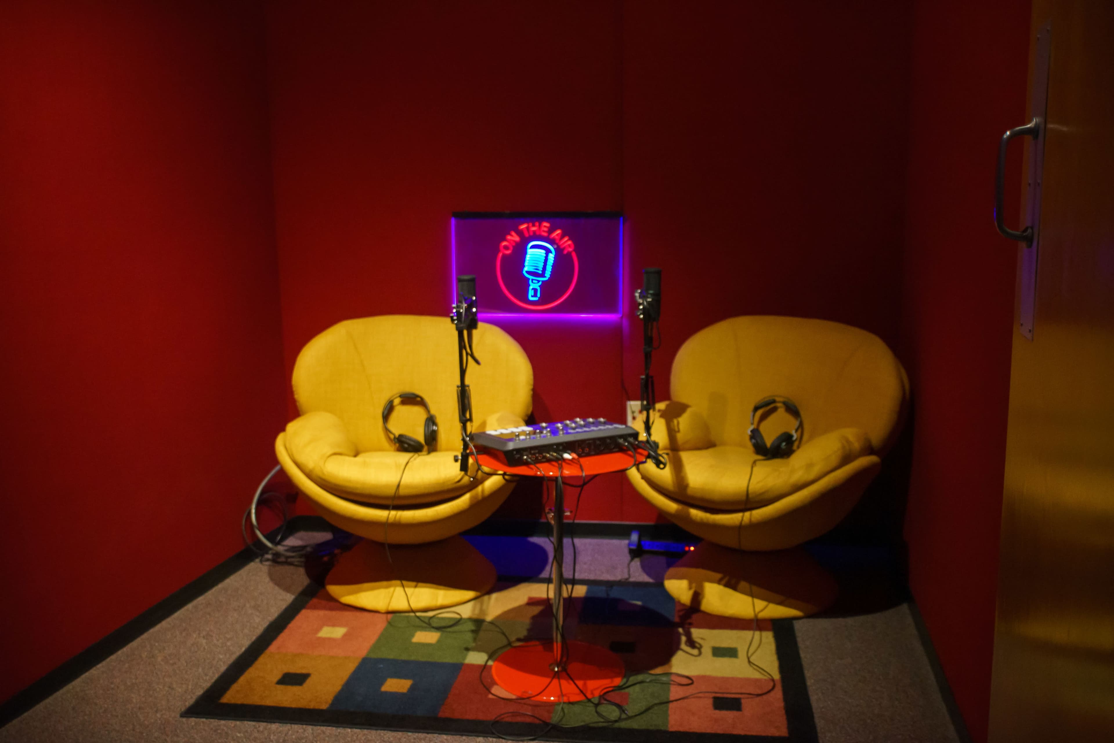 A small recording studio features two yellow chairs, microphones, and a musical mixing table against a red backdrop.