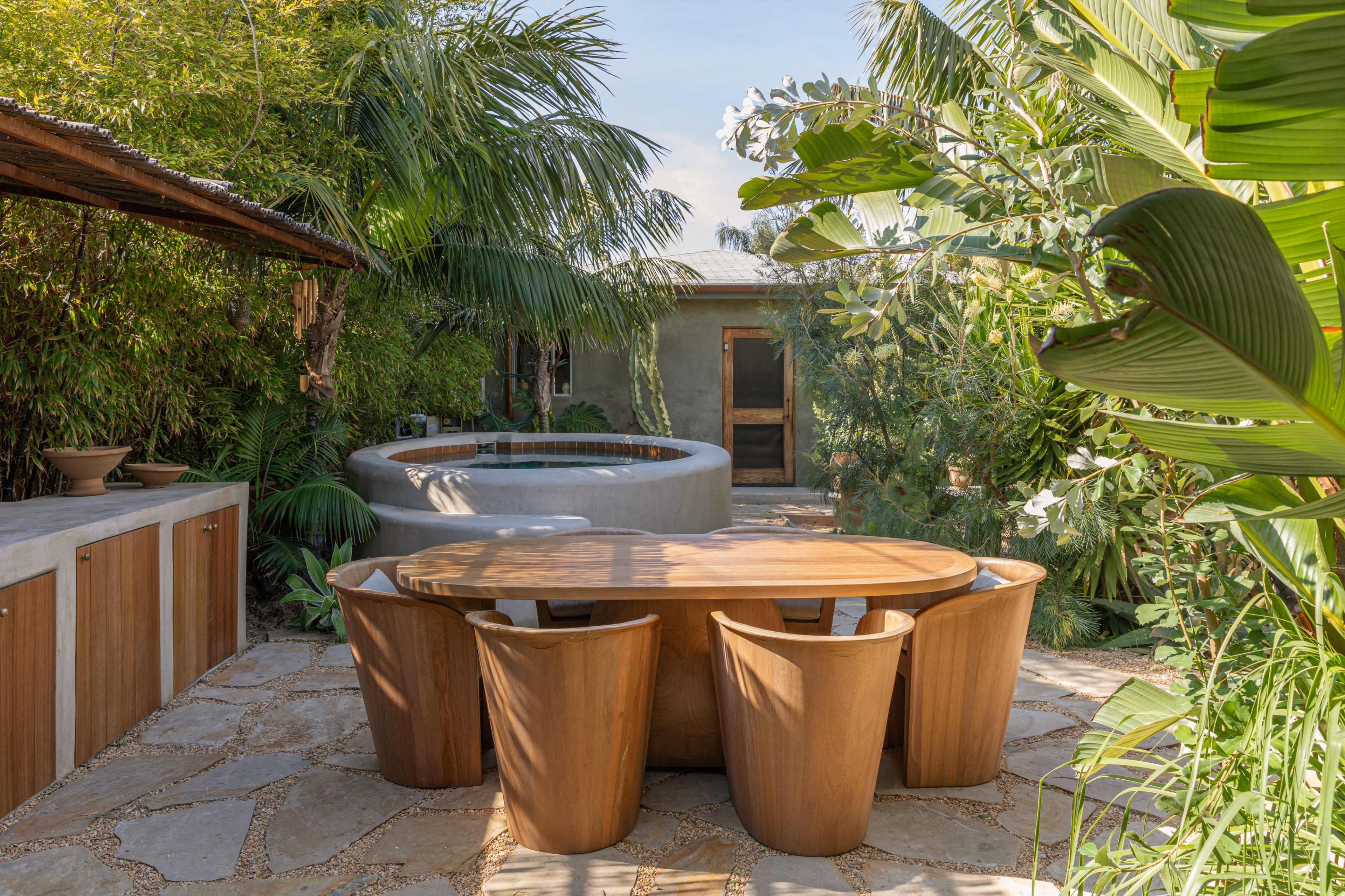 A wooden dining table surrounded by shaped chairs is set in a garden with lush greenery and a circular hot tub nearby.
