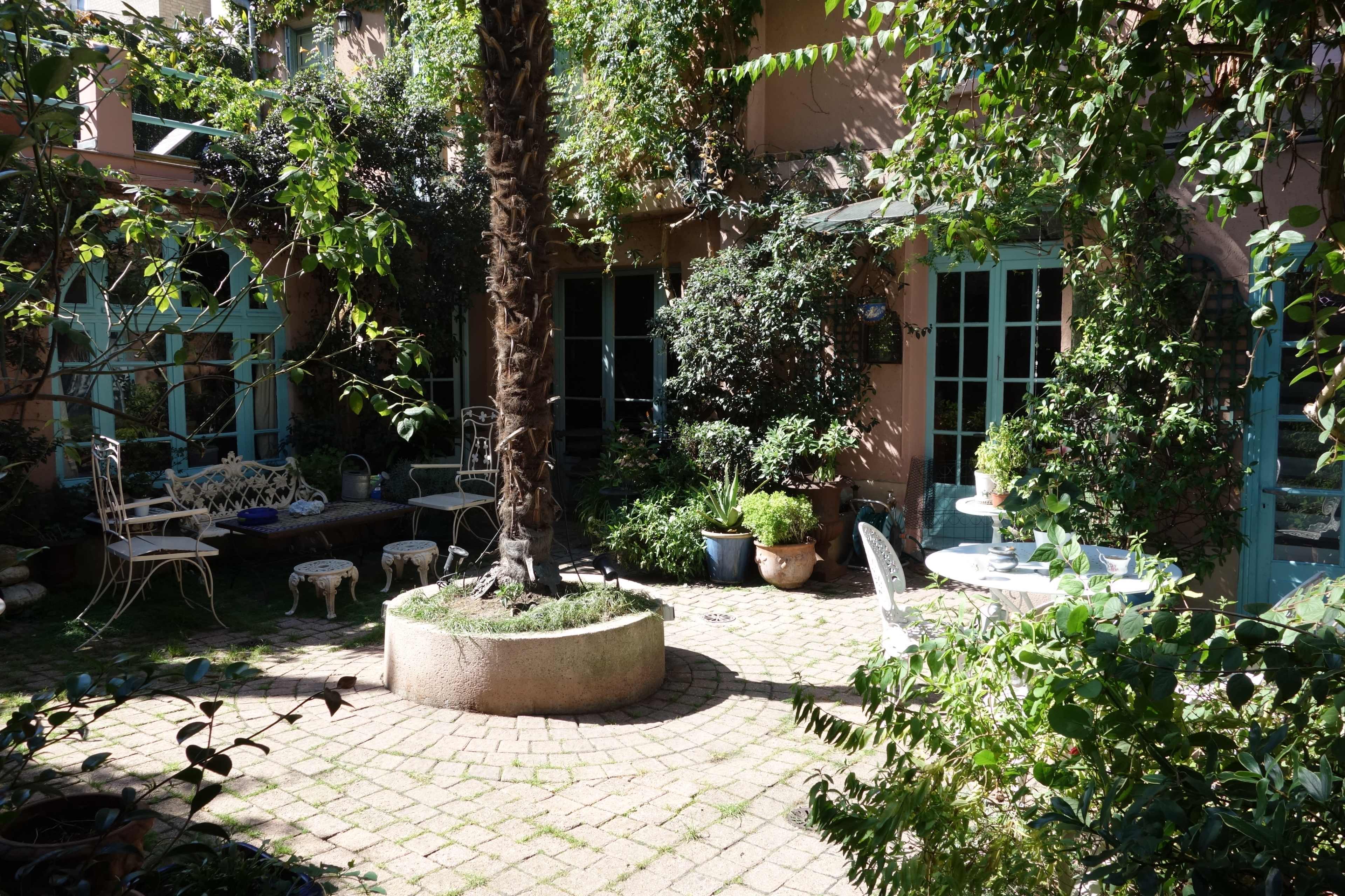 The scene features a lush courtyard with stone-paved paths, greenery, and outdoor seating arrangements surrounded by walls adorned with plants.