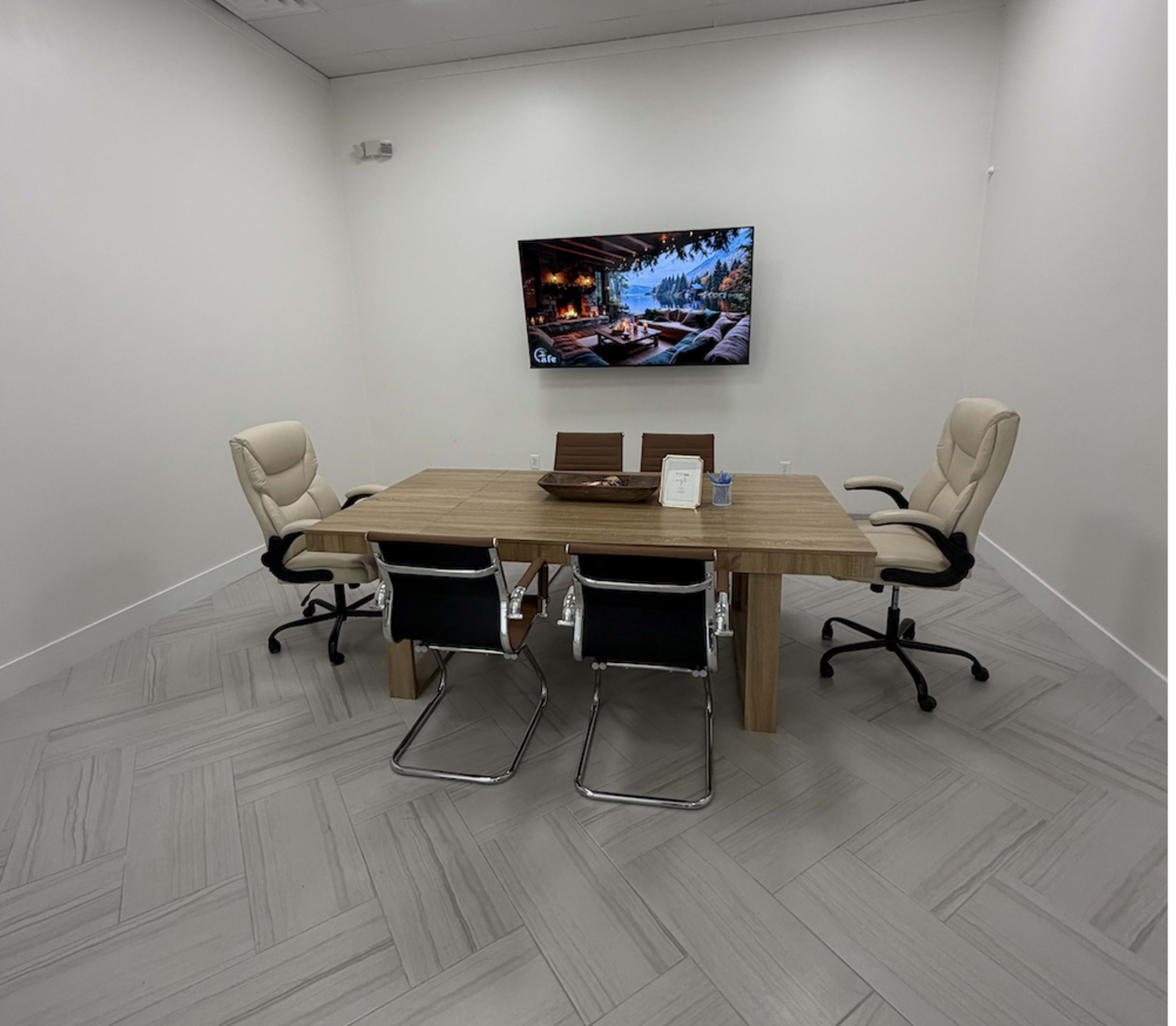 A small, modern conference room features a wooden table with four chairs and a wall-mounted TV displaying a scenic image.