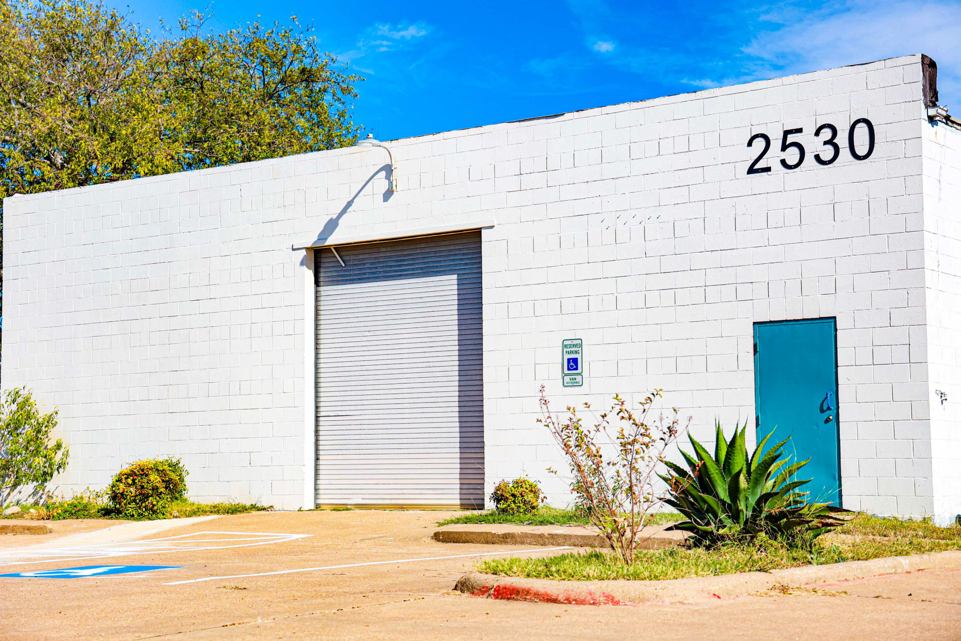 A single-story white building with a large silver roll-up door and a blue door, located at 2530, surrounded by a small parking area and some greenery.