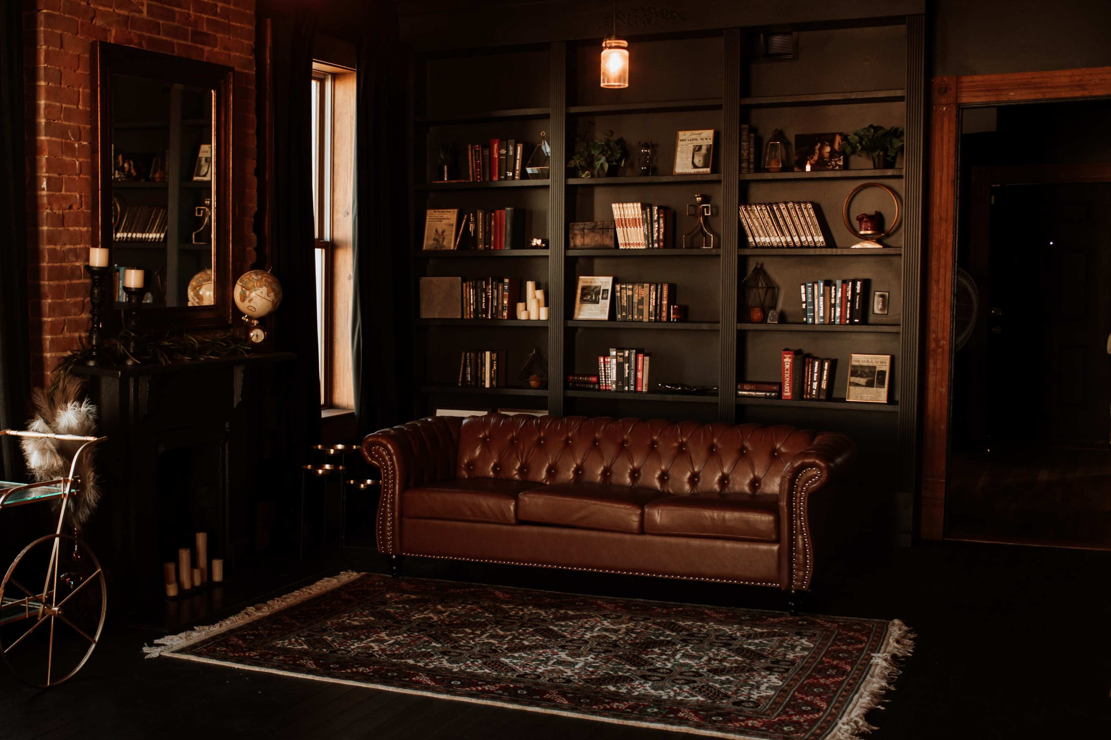 A cozy living room featuring a brown leather sofa, a dark bookshelf filled with books and decorative items, and a fireplace with candles.
