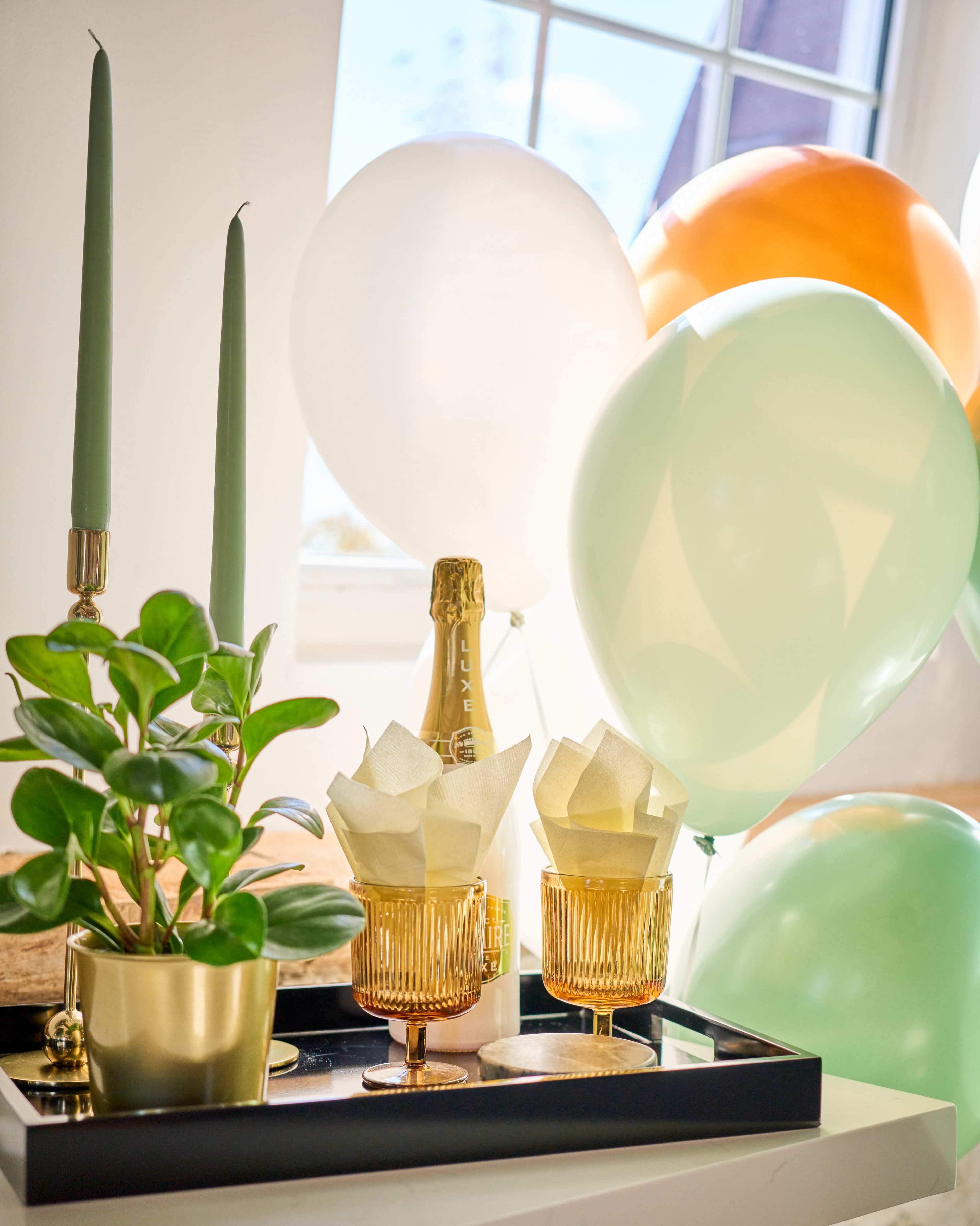 A decorative tray holds two golden glasses with neatly folded napkins, a small potted plant, a bottle of champagne, and colorful balloons in a brightly lit room.