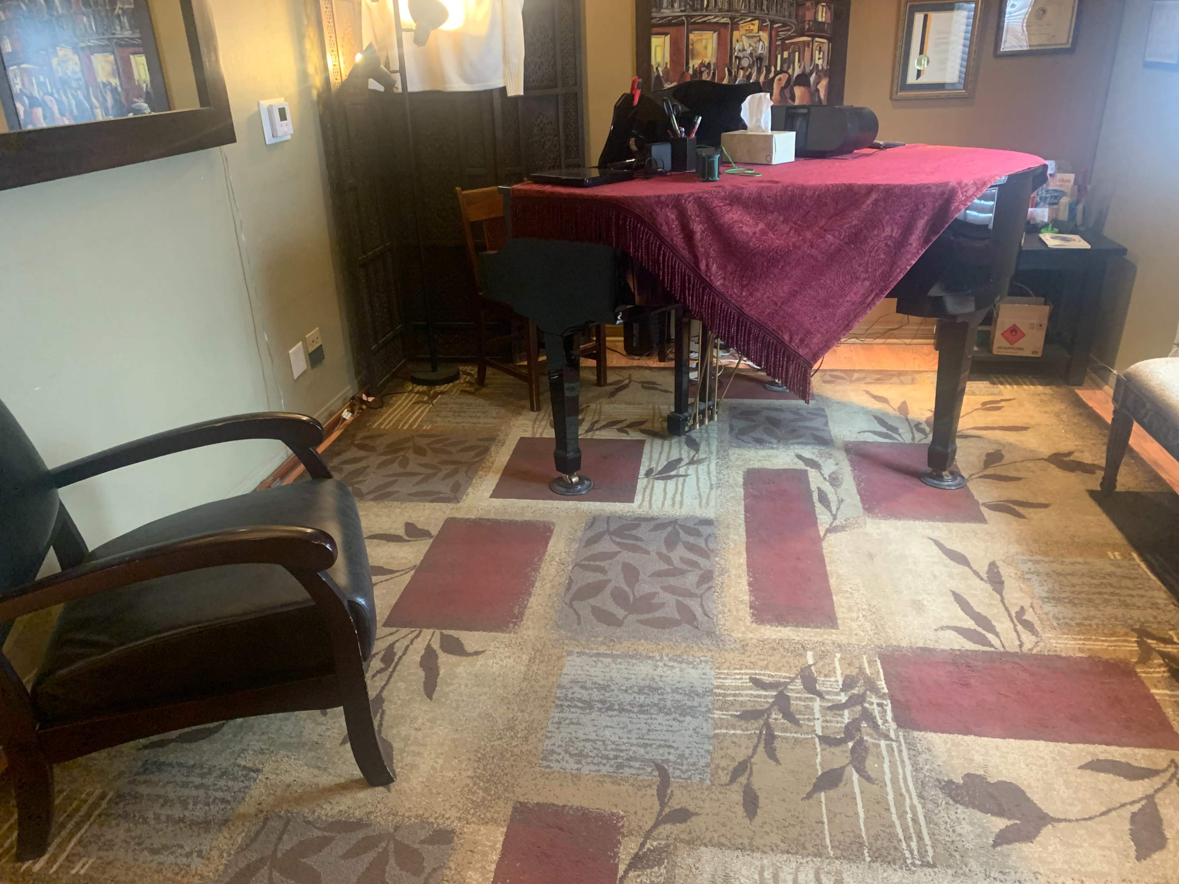 A grand piano covered with a maroon cloth sits in a room featuring a patterned carpet and a black chair nearby.