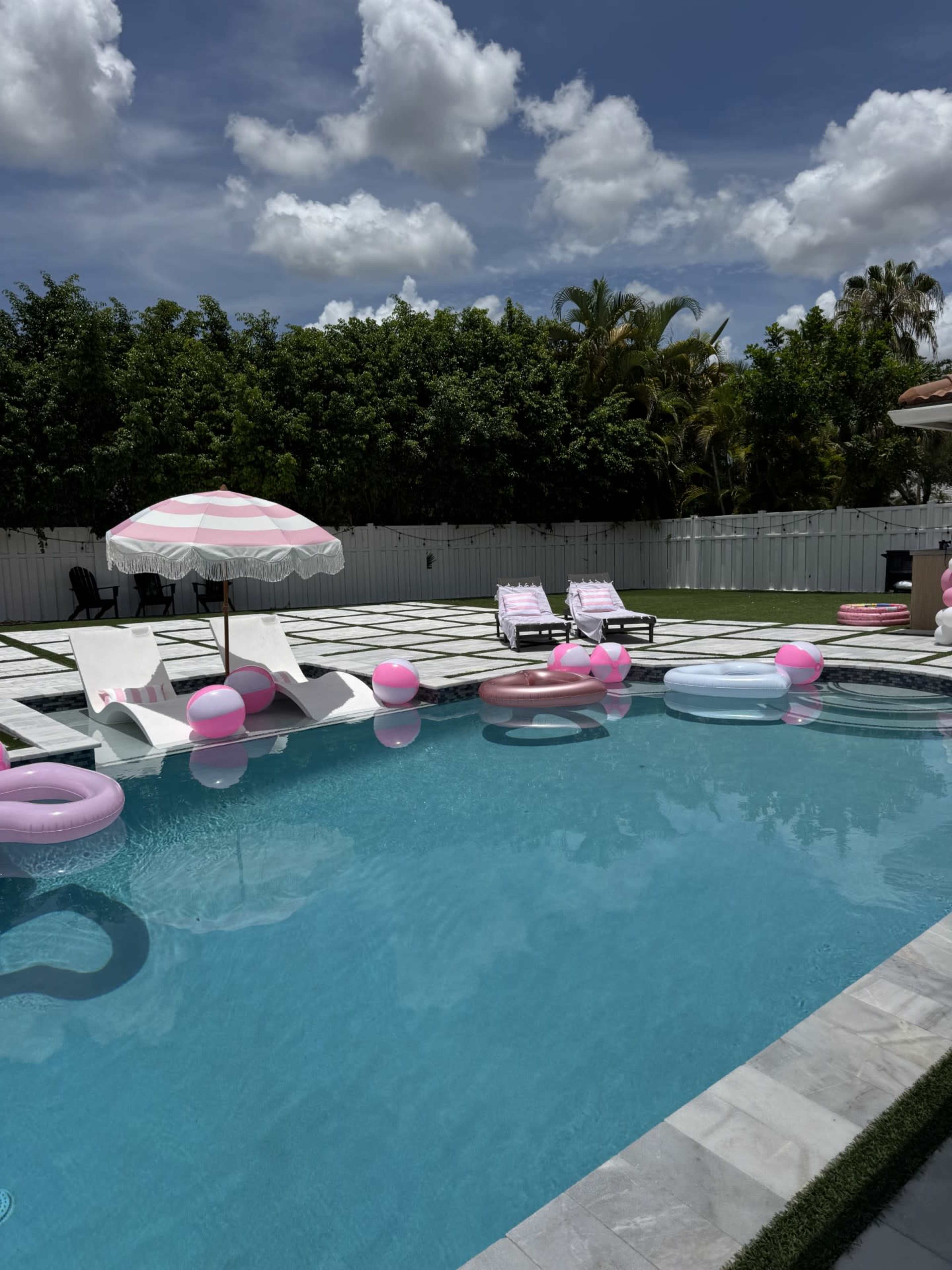 A backyard swimming pool features colorful inflatable floats, lounge chairs, and an umbrella under a partly cloudy sky.