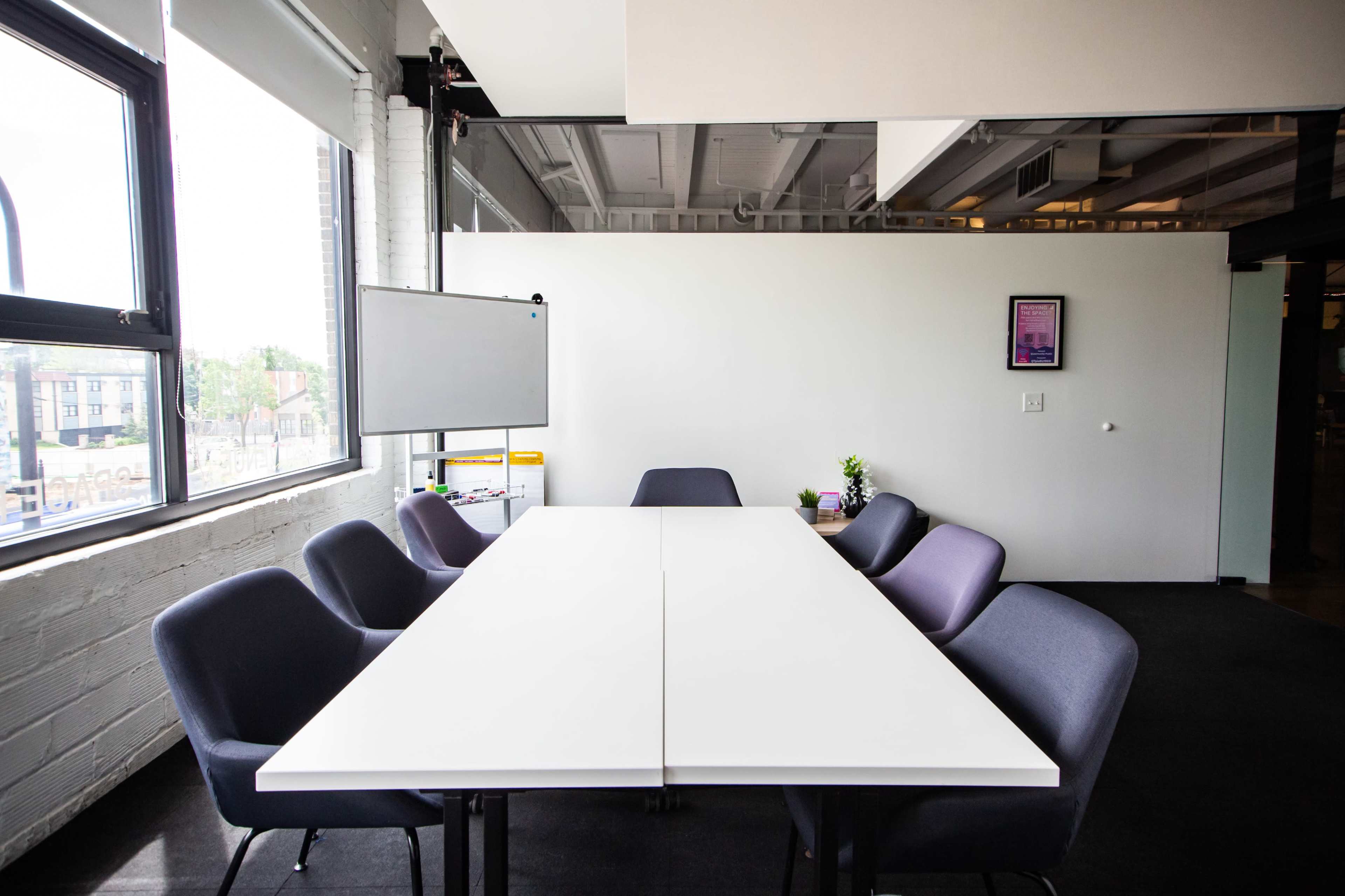 A long, white conference table is surrounded by gray and purple chairs in a well-lit meeting room with large windows.
