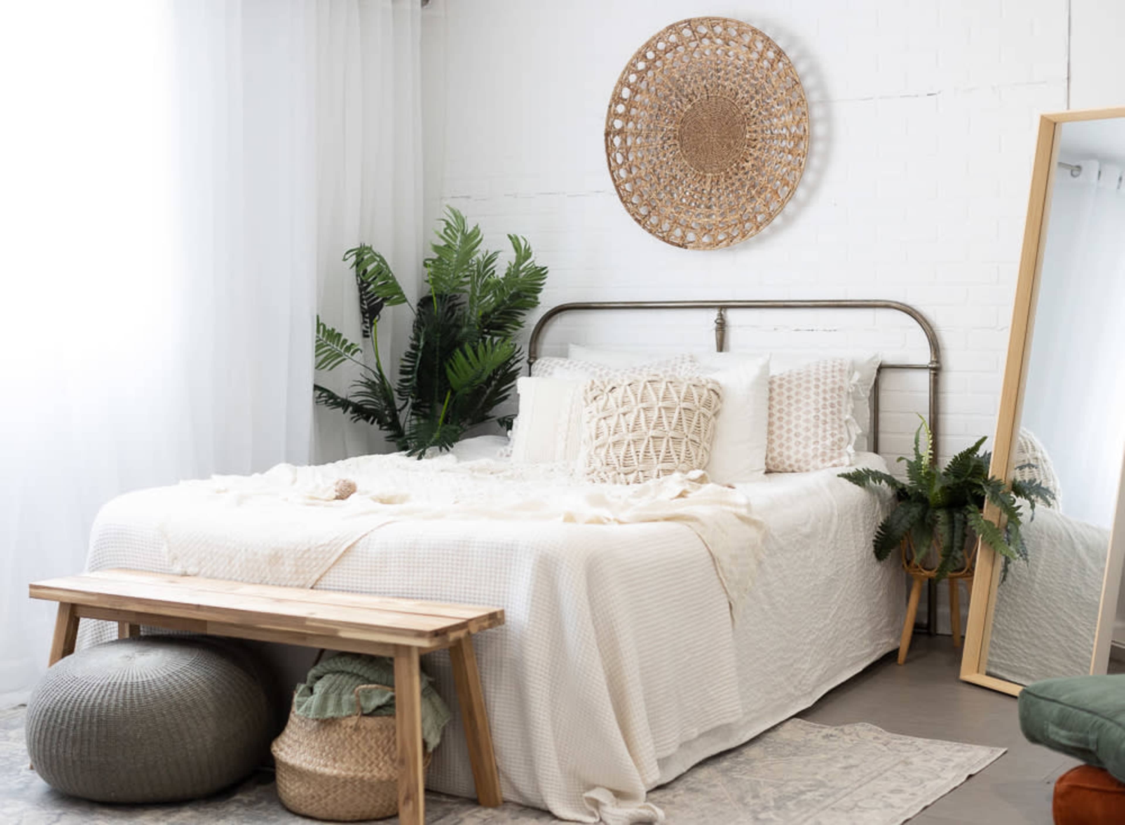 The scene shows a tidy bedroom with a bed dressed in neutral linens, a decorative wall hanging, potted plants, a large mirror, and a wooden bench at the foot of the bed.